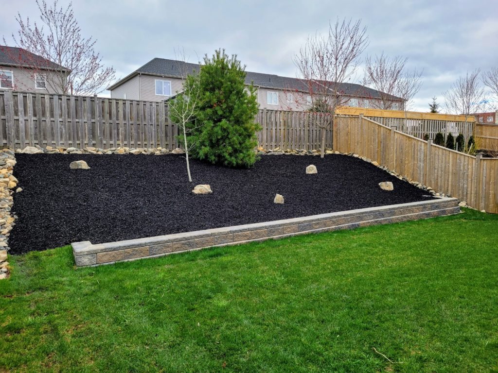 A backyard features a tiered garden with dark mulch, rocks, and a small tree, surrounded by wooden fencing and grass.