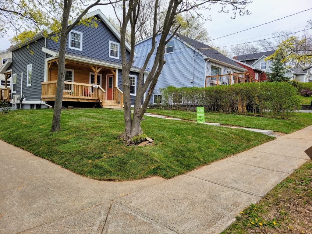 A charming, blue-gray house with a porch is surrounded by lush greenery and a sidewalk, featuring a small sign on the lawn.