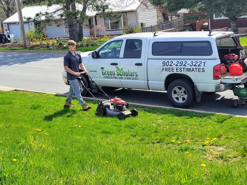 A person mowing a lawn near a truck labeled "Green Scholars Landscaping and Lawn Care" parked on a residential street.