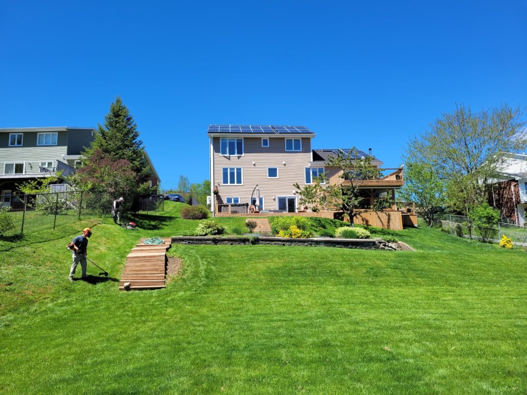 A house with solar panels and a wooden deck overlooks a green lawn. Two people are gardening under a clear blue sky.