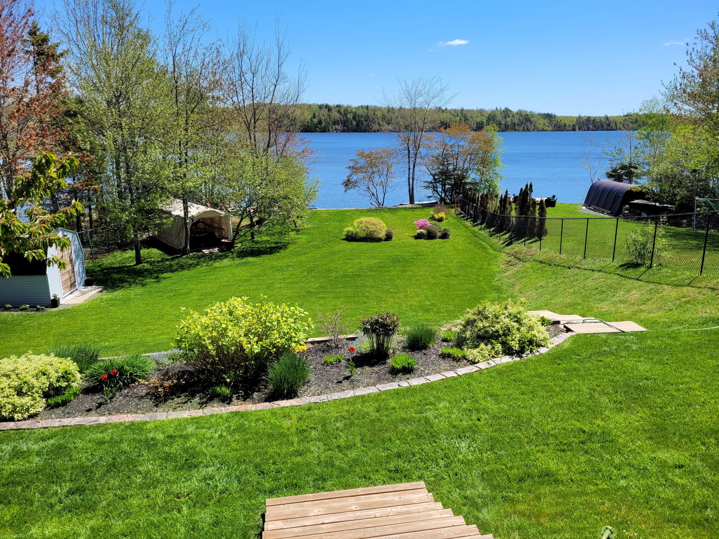 A serene lakeside backyard with lush greenery, a small shed, and a canopy beside the clear blue water under a sunny sky.