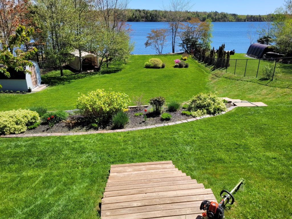 A well-maintained backyard garden with a wooden deck, lake view, and surrounding trees on a sunny day. A shed and tent are visible.