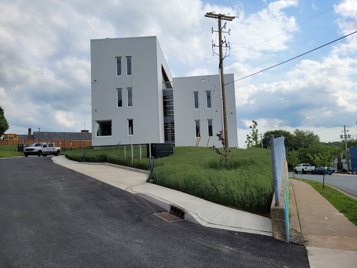 A modern white building on a grassy hill, with a utility pole nearby and parked vehicles on the road under a cloudy sky.