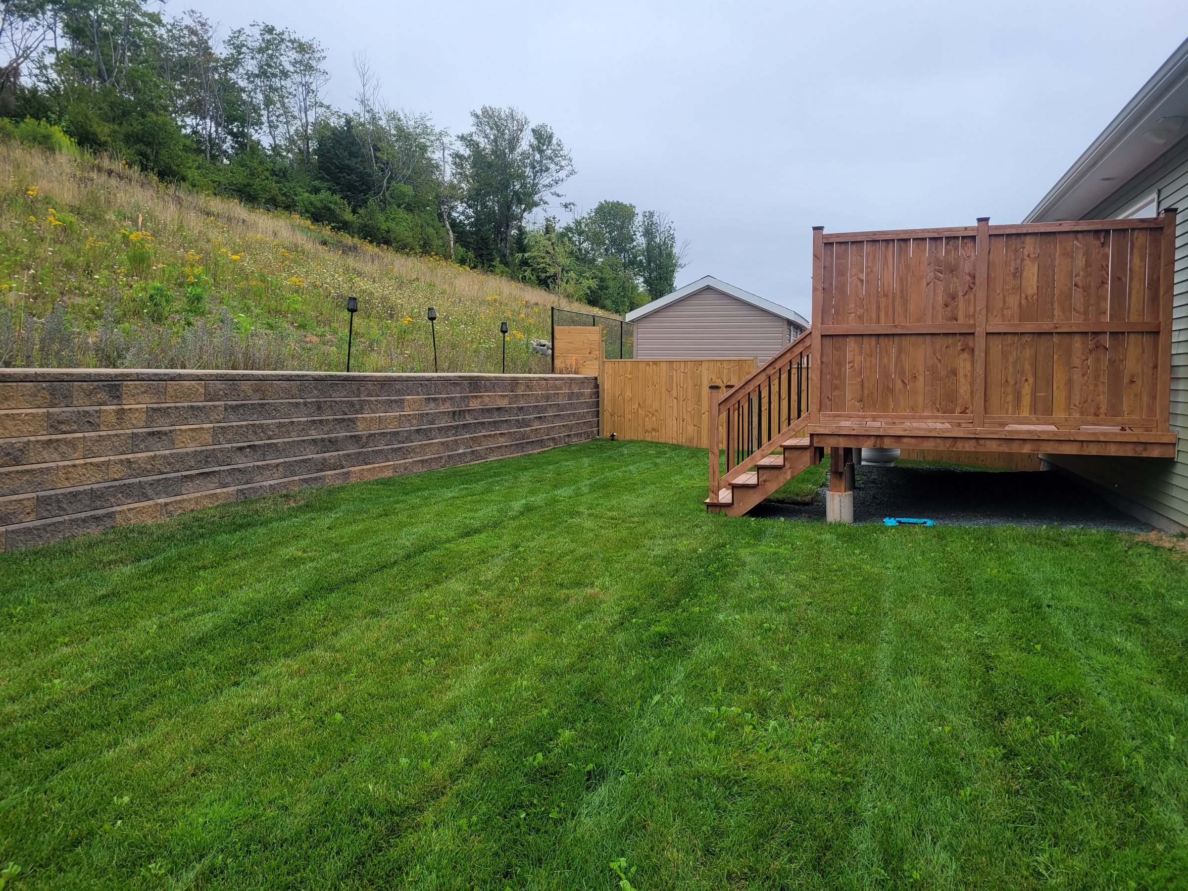 A wooden deck with stairs overlooks a lush green backyard, bordered by a retaining wall and fence, adjacent to a hillside.