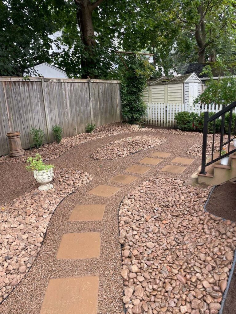 A neatly designed garden path with stepping stones, surrounded by gravel, trees, and a wooden fence, leads to a small shed.