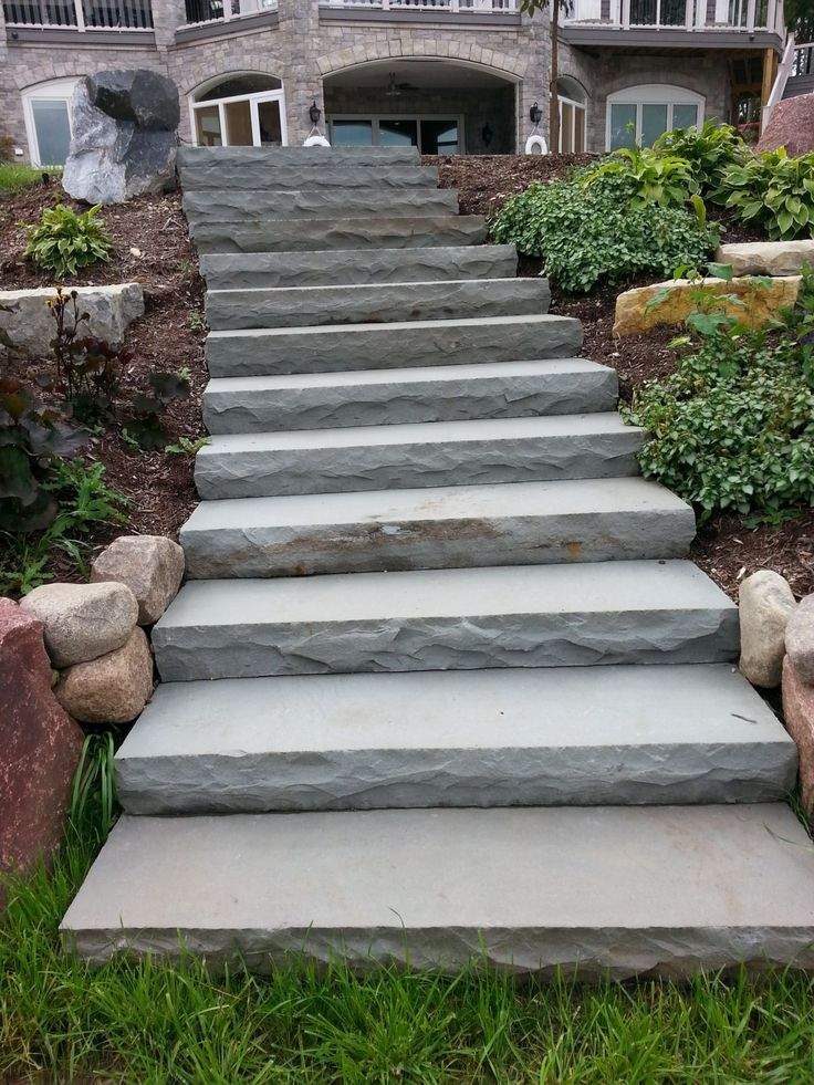 Stone steps lead up to a large, elegant residential building, surrounded by green plants and rocks, creating a natural, inviting entrance.