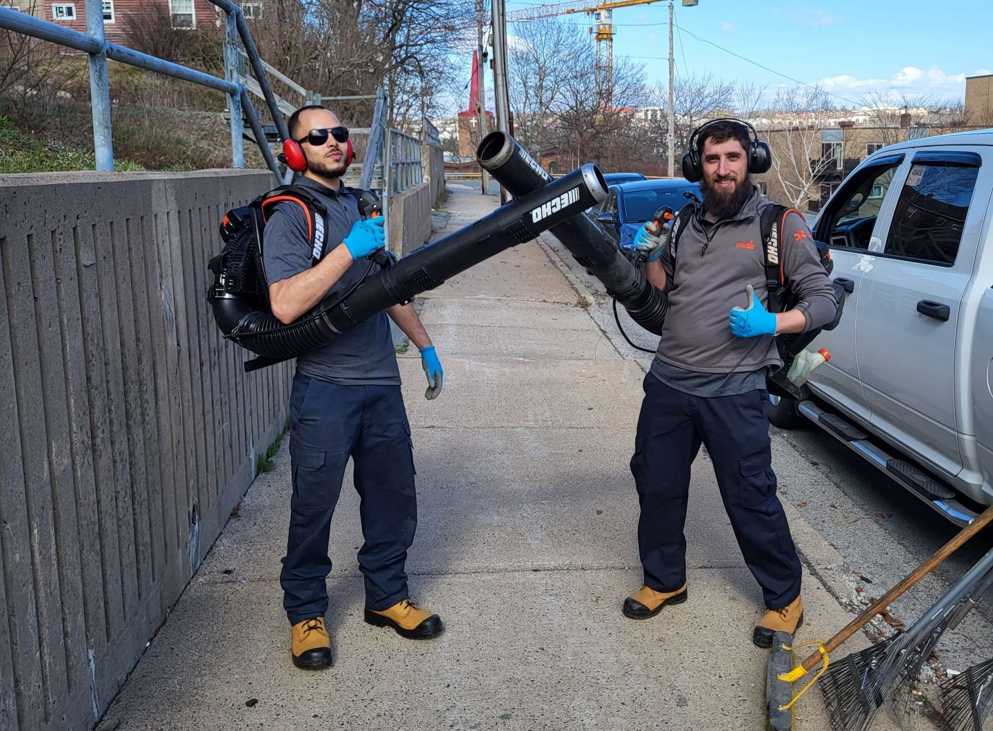 Two people with leaf blowers strike playful poses on a sidewalk, wearing safety gear. Construction crane visible in the background.