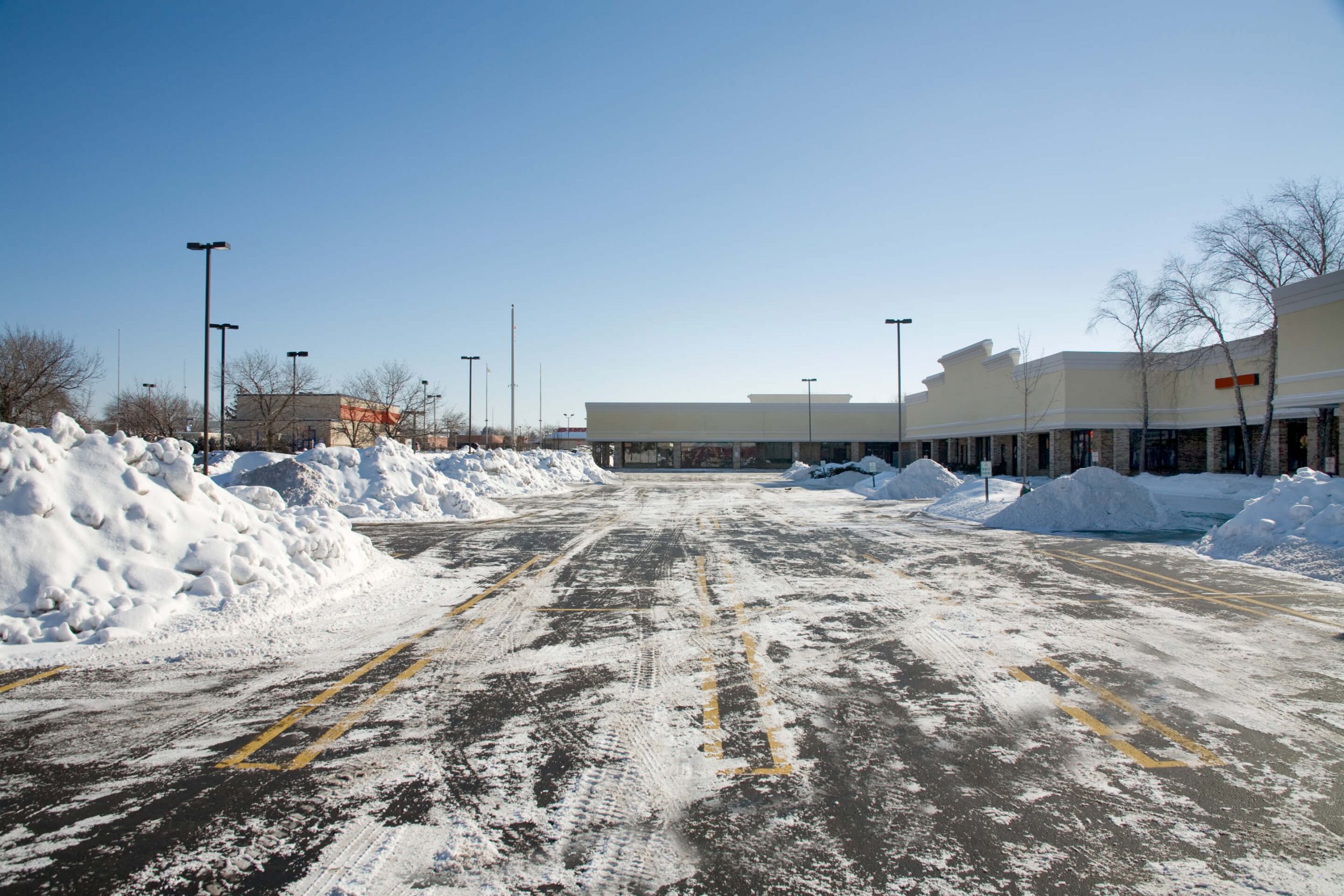 Snow-covered, empty parking lot with plowed snow piles, surrounded by a strip mall under a clear blue sky, no recognizable landmarks present.