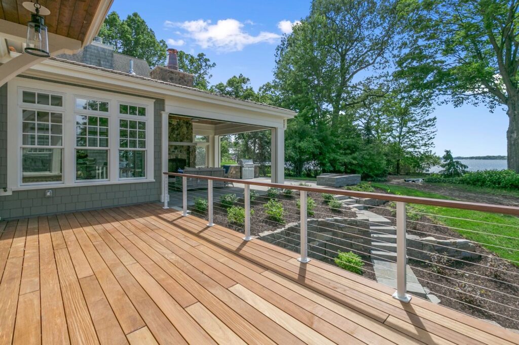 Spacious wooden deck overlooking a garden with stone steps, surrounded by trees and water, under a clear blue sky.