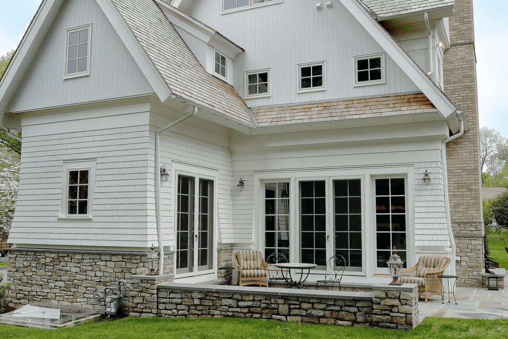 A large, modern white house with stone accents, featuring a patio with chairs and table, surrounded by green grass and trees.