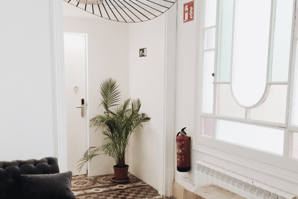 Cozy interior with a corner plant, stylish lamp, patterned floor, and white door. A fire extinguisher hangs near a stained glass window.