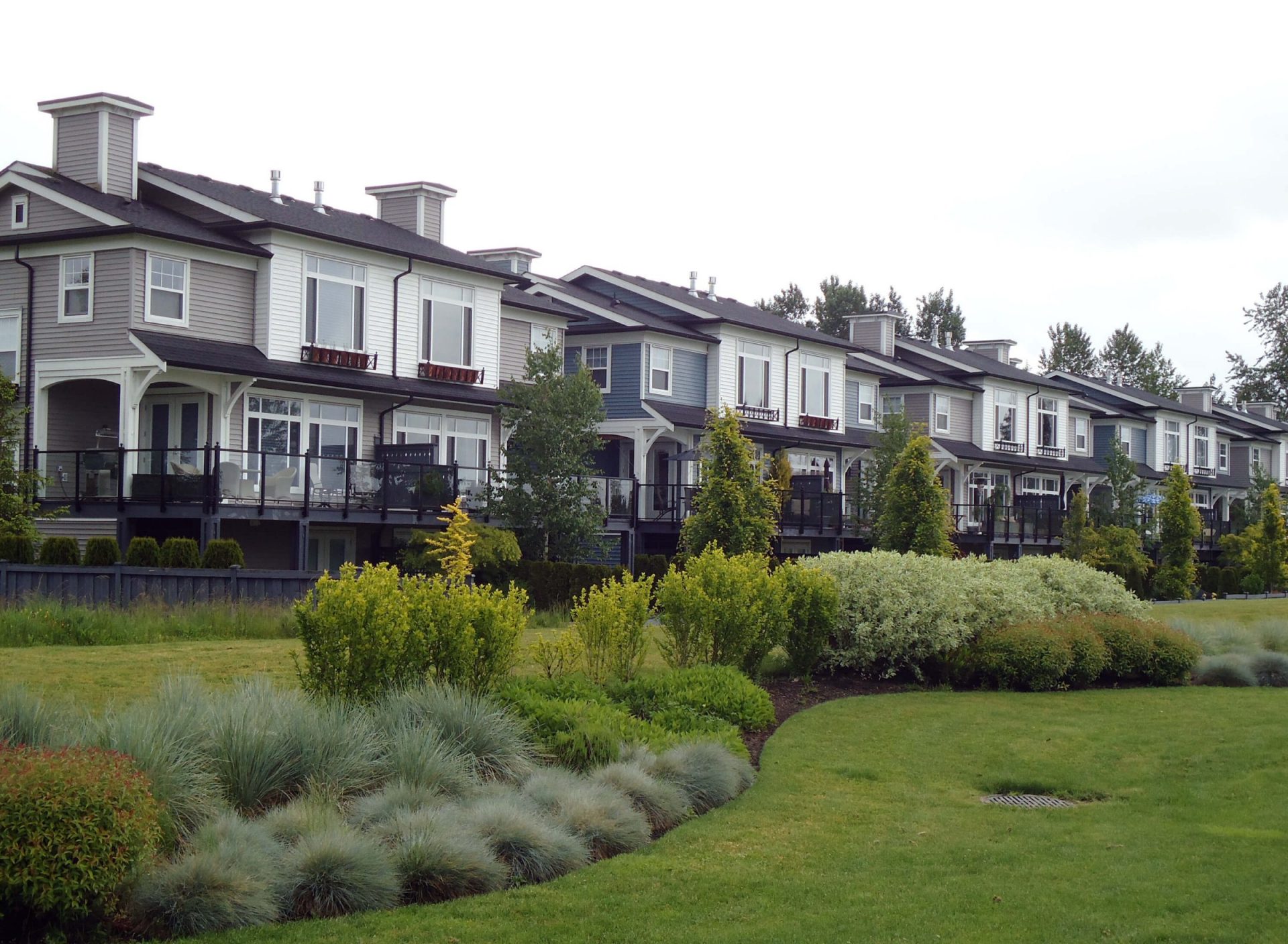A row of modern townhouses with white and gray exteriors, bordered by landscaped gardens and lush greenery under a cloudy sky.