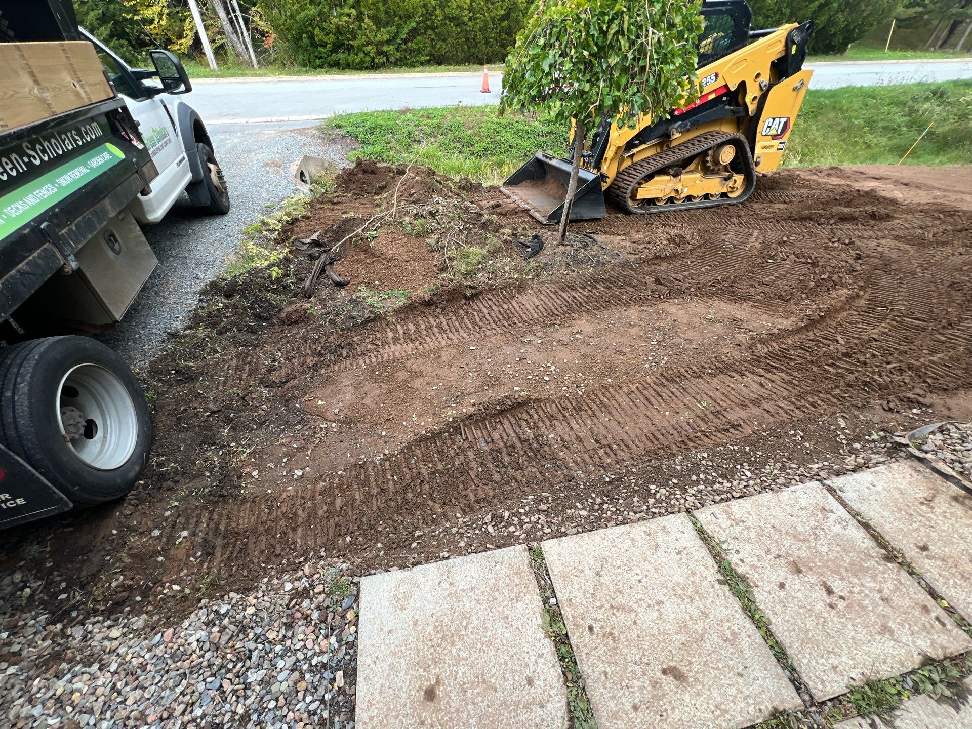 A small construction site with a skid steer loader and a parked truck. Dirt and gravel cover the ground near a paved walkway.