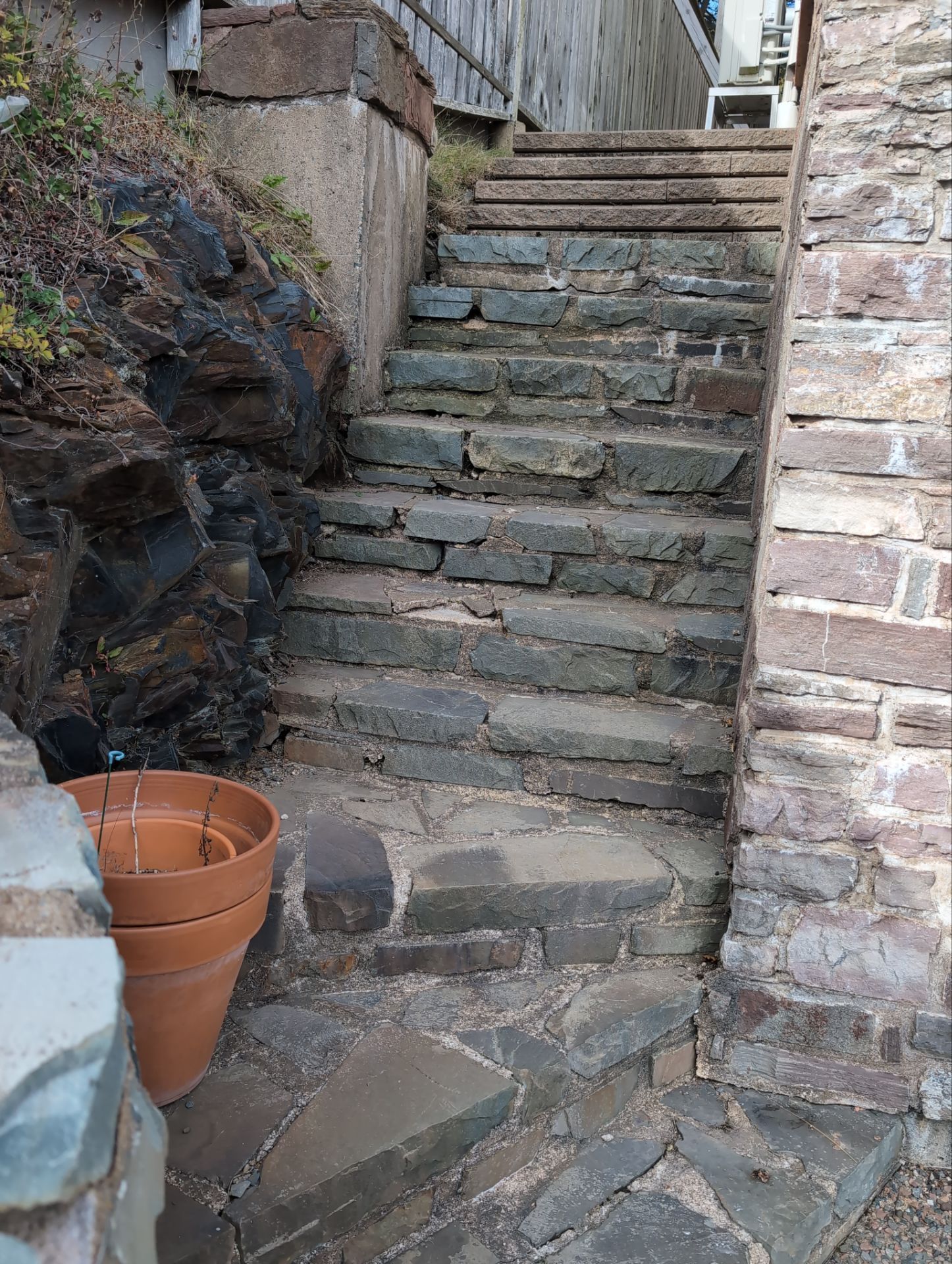 Stone steps leading upward, flanked by a wooden fence and rock wall. A terra cotta pot sits on the steps in natural surroundings.