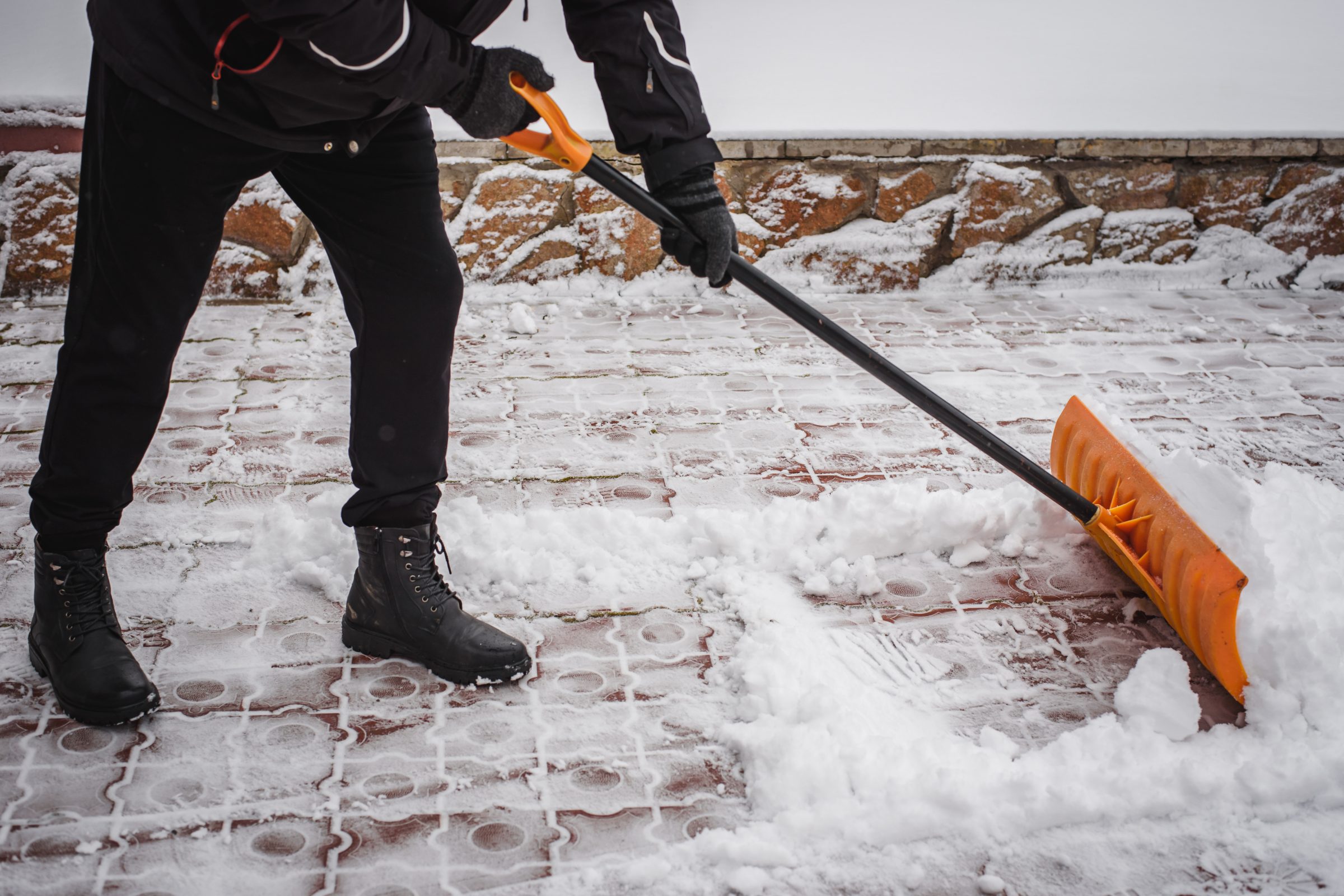 A person in black clothing shovels snow from a brick path, using an orange snow shovel, with a stone wall in the background.