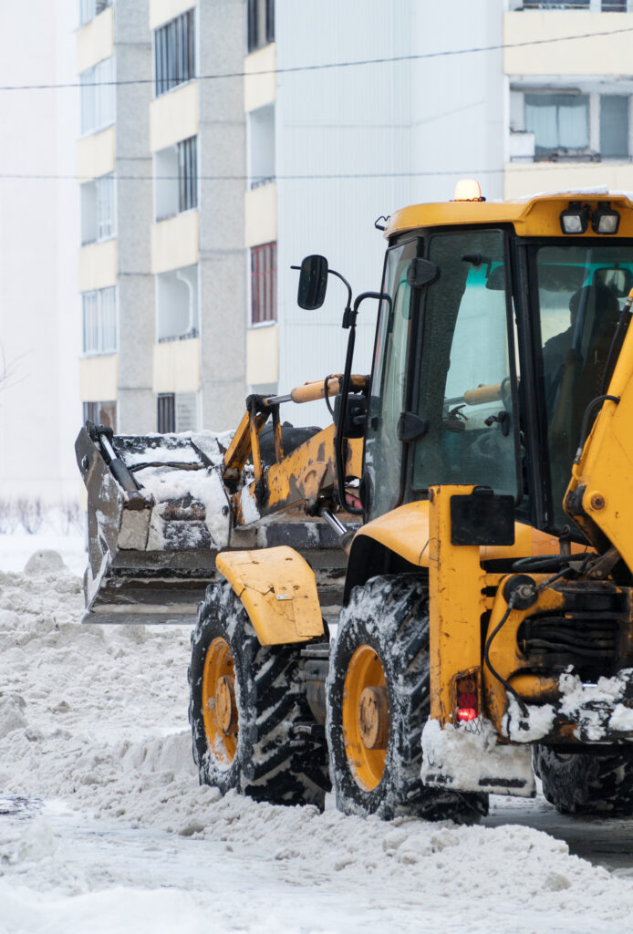 A yellow snowplow clearing snow on a street, surrounded by apartment buildings. Overcast weather and accumulated snow suggest a cold, wintry day.