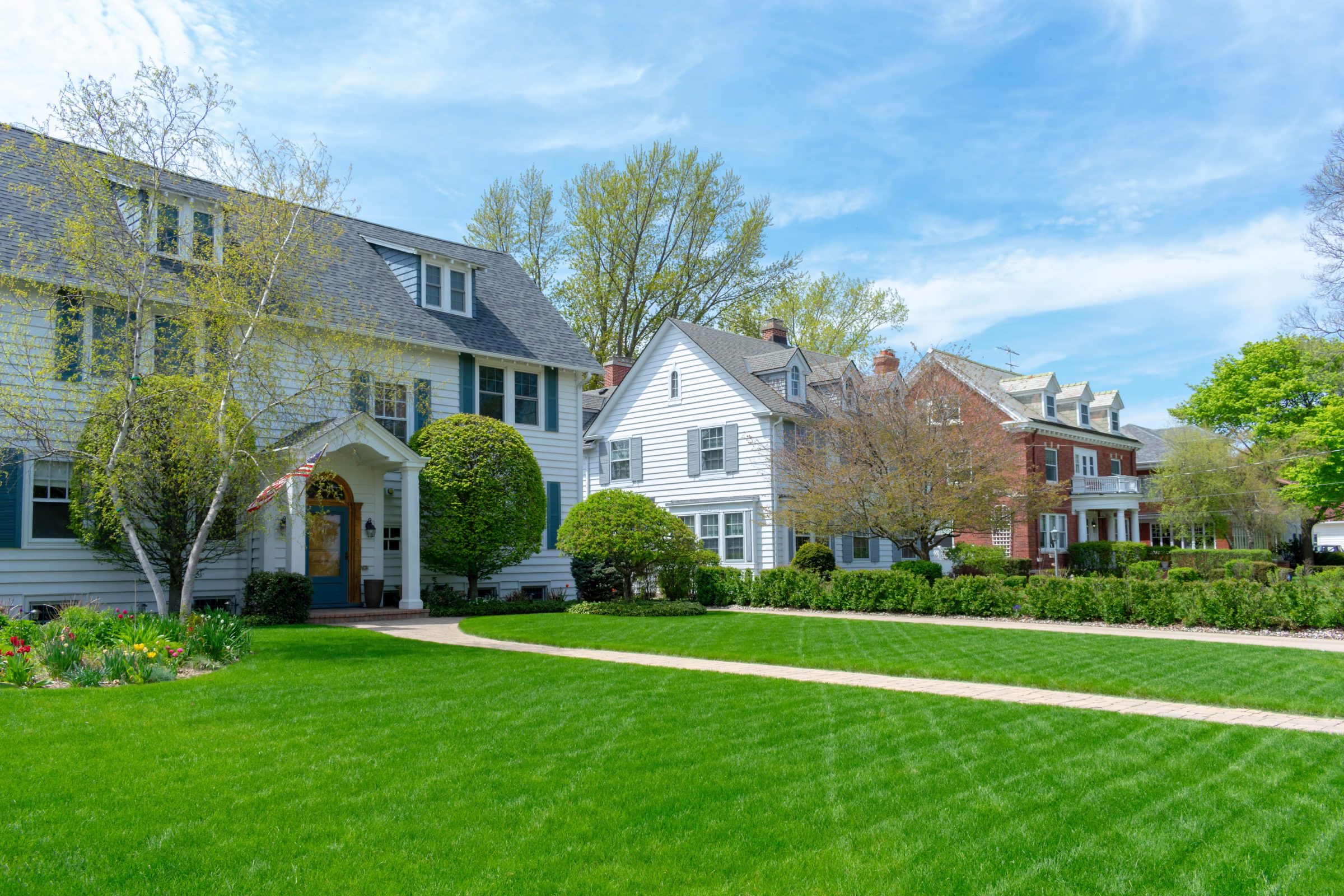 Traditional suburban neighborhood with well-maintained lawns, trees, and charming houses with different architectural styles under a clear blue sky.