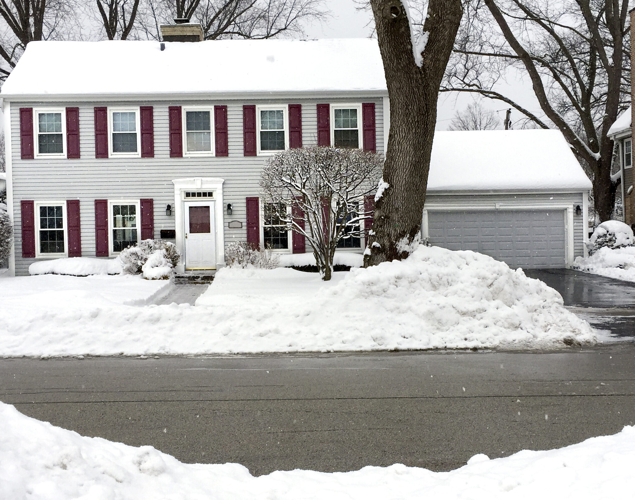 A snow-covered suburban house with red shutters, surrounded by trees and a driveway. Snow blankets the yard and street.
