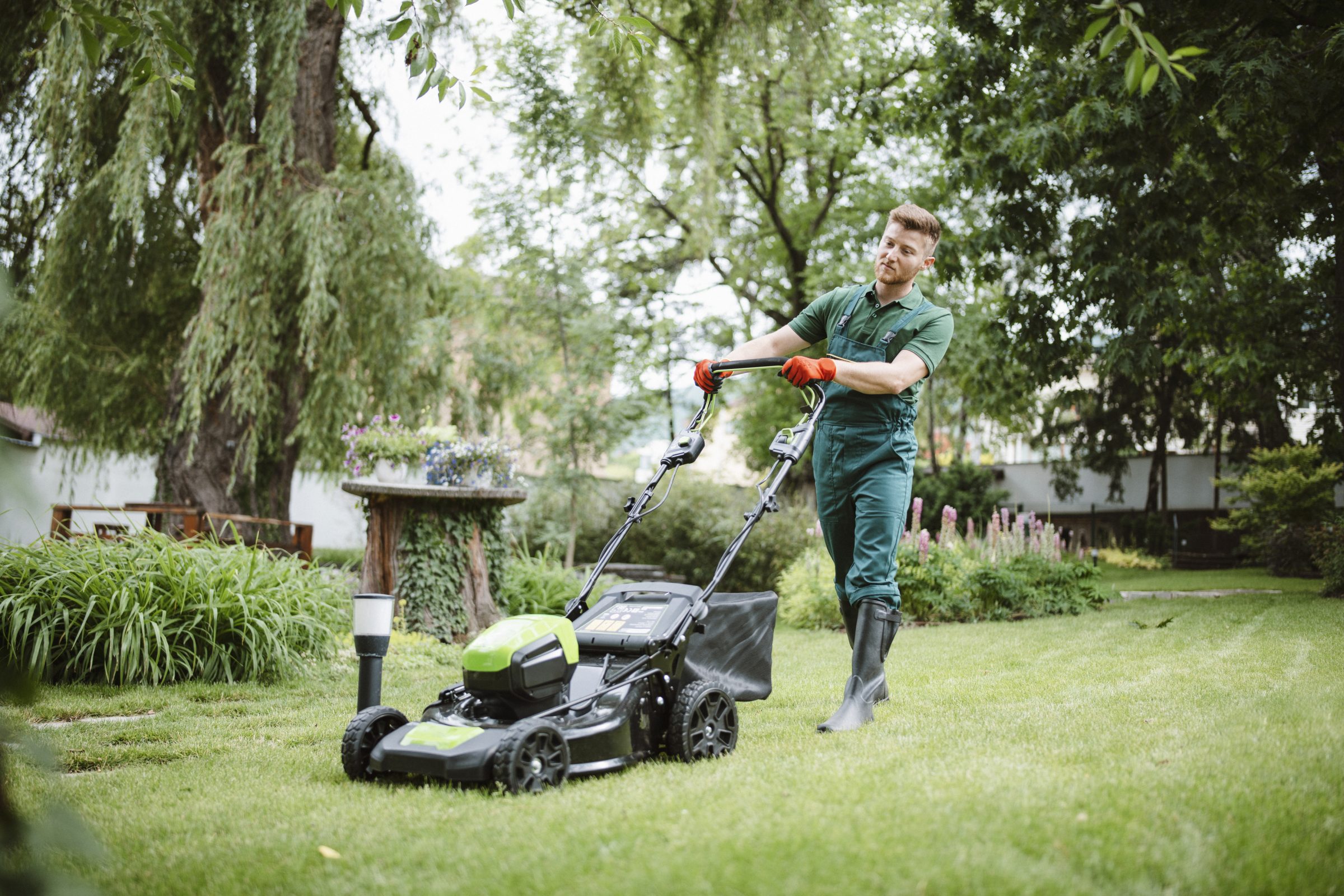 A person in green overalls cutting grass with a lawnmower in a lush garden, surrounded by trees and plants.