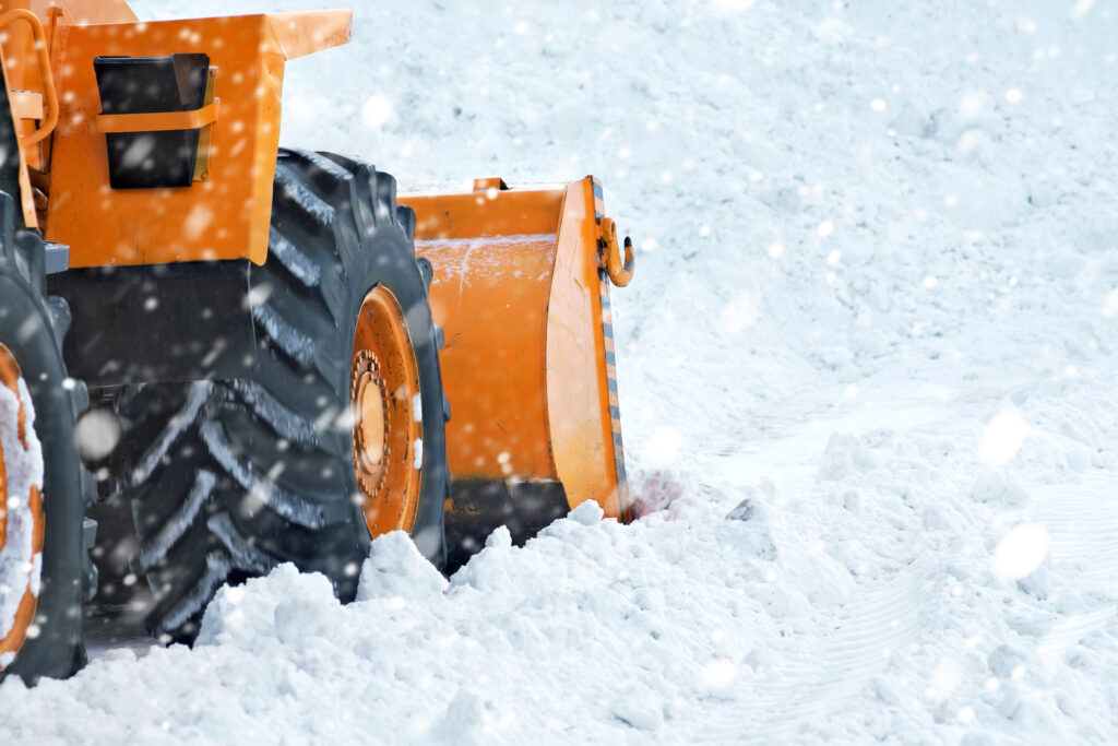 An orange snowplow pushes through a thick layer of snow. Snowflakes fall around the machine in an active winter scene.
