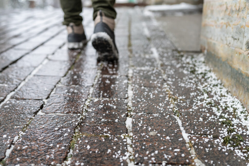 A person walks on a wet, snow-covered brick sidewalk; legs in view. The wall next to them is slightly textured and worn.