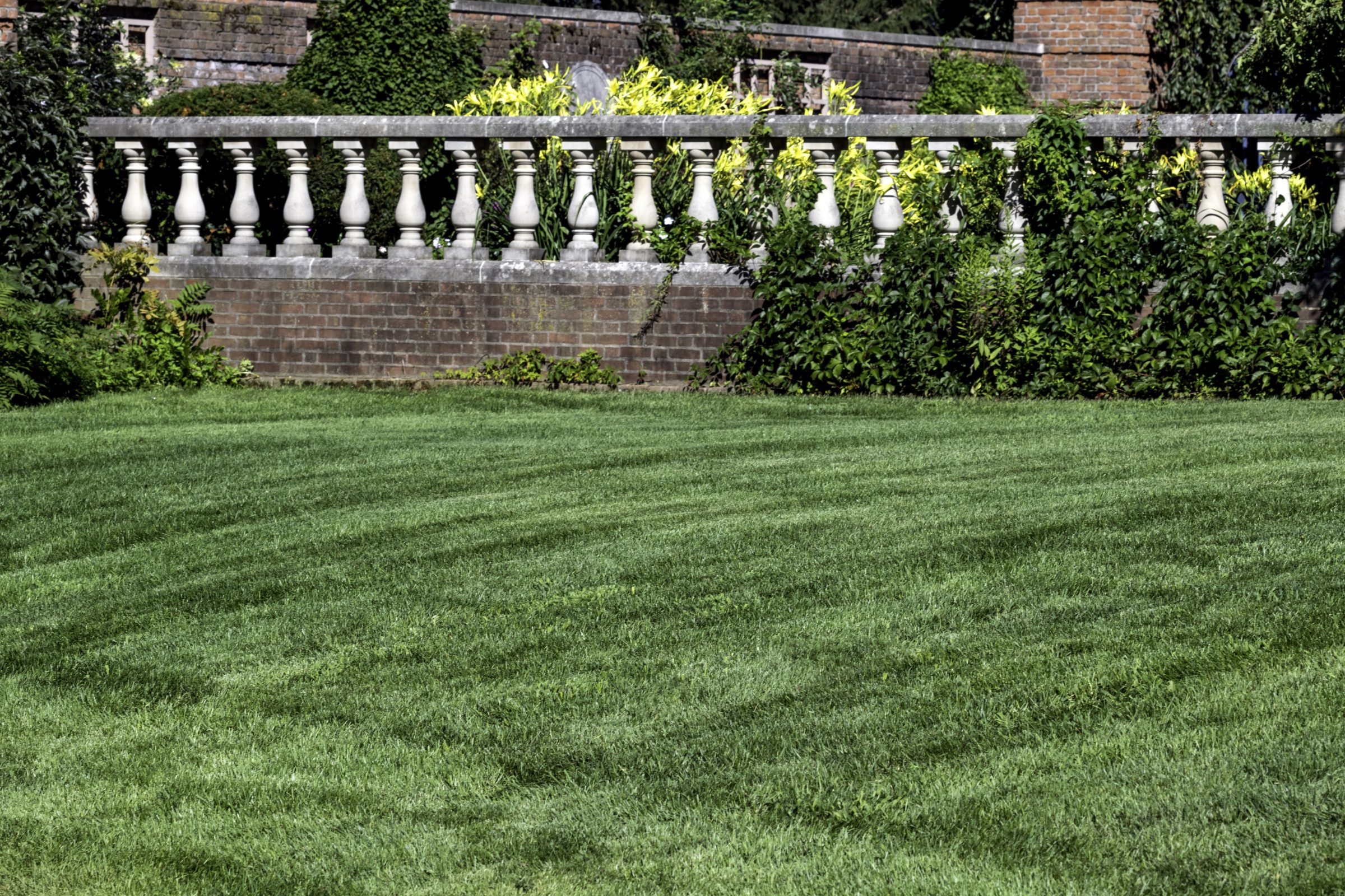 A well-maintained grassy lawn leads to a decorative stone balustrade, surrounded by lush plants and greenery, against a brick wall backdrop.