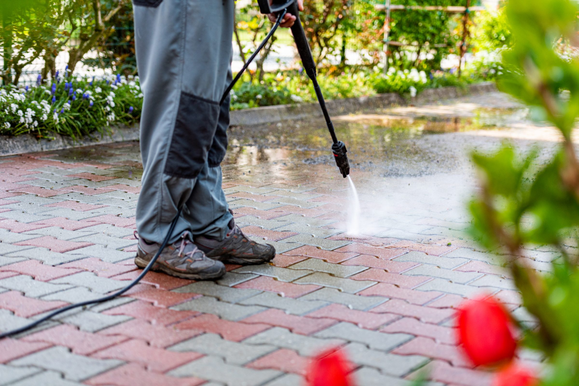A person uses a pressure washer to clean a patterned brick driveway, surrounded by greenery and vibrant flowers.