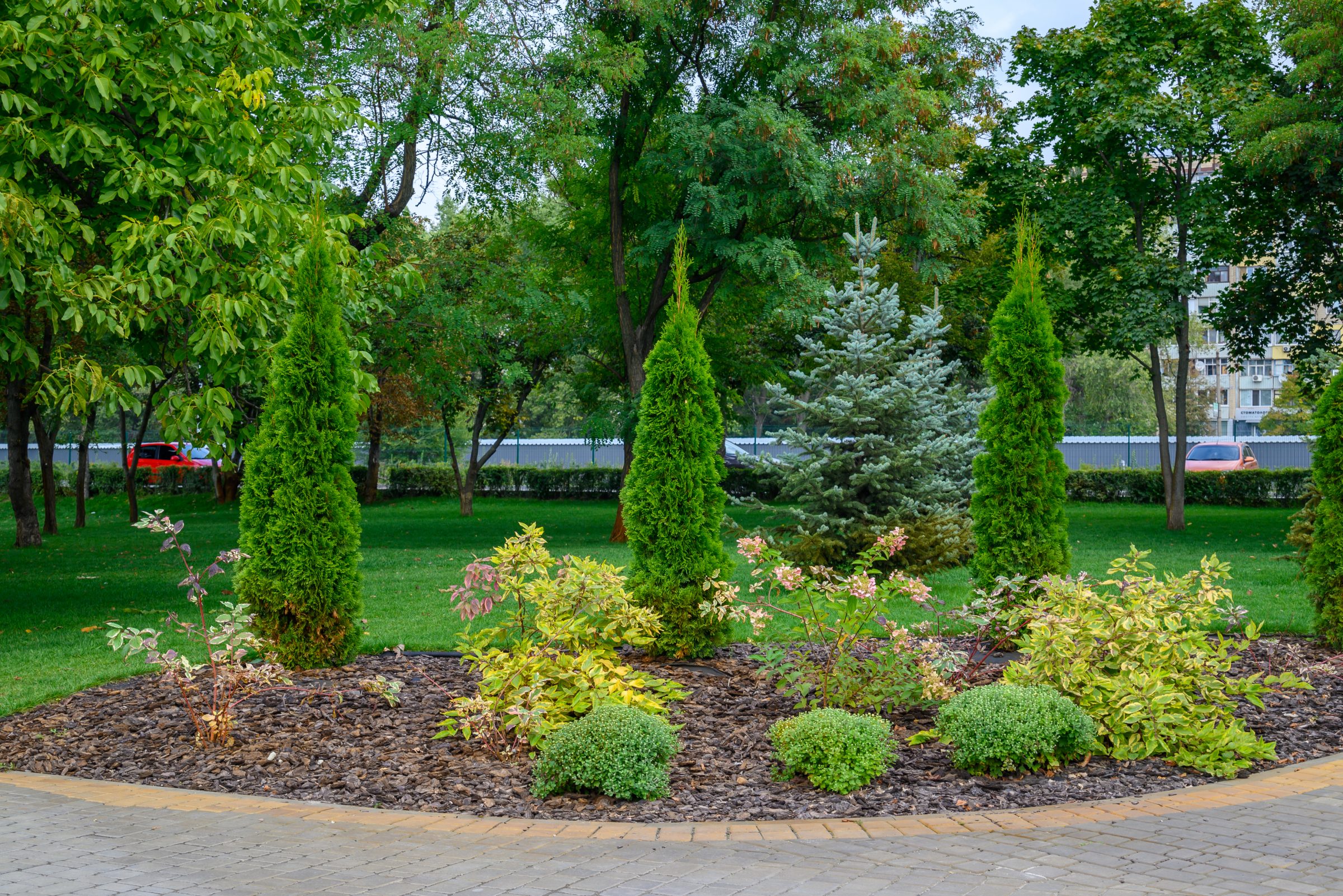 A landscaped park area with neatly trimmed bushes and trees, bordered by a paved path, with residential buildings in the backdrop.