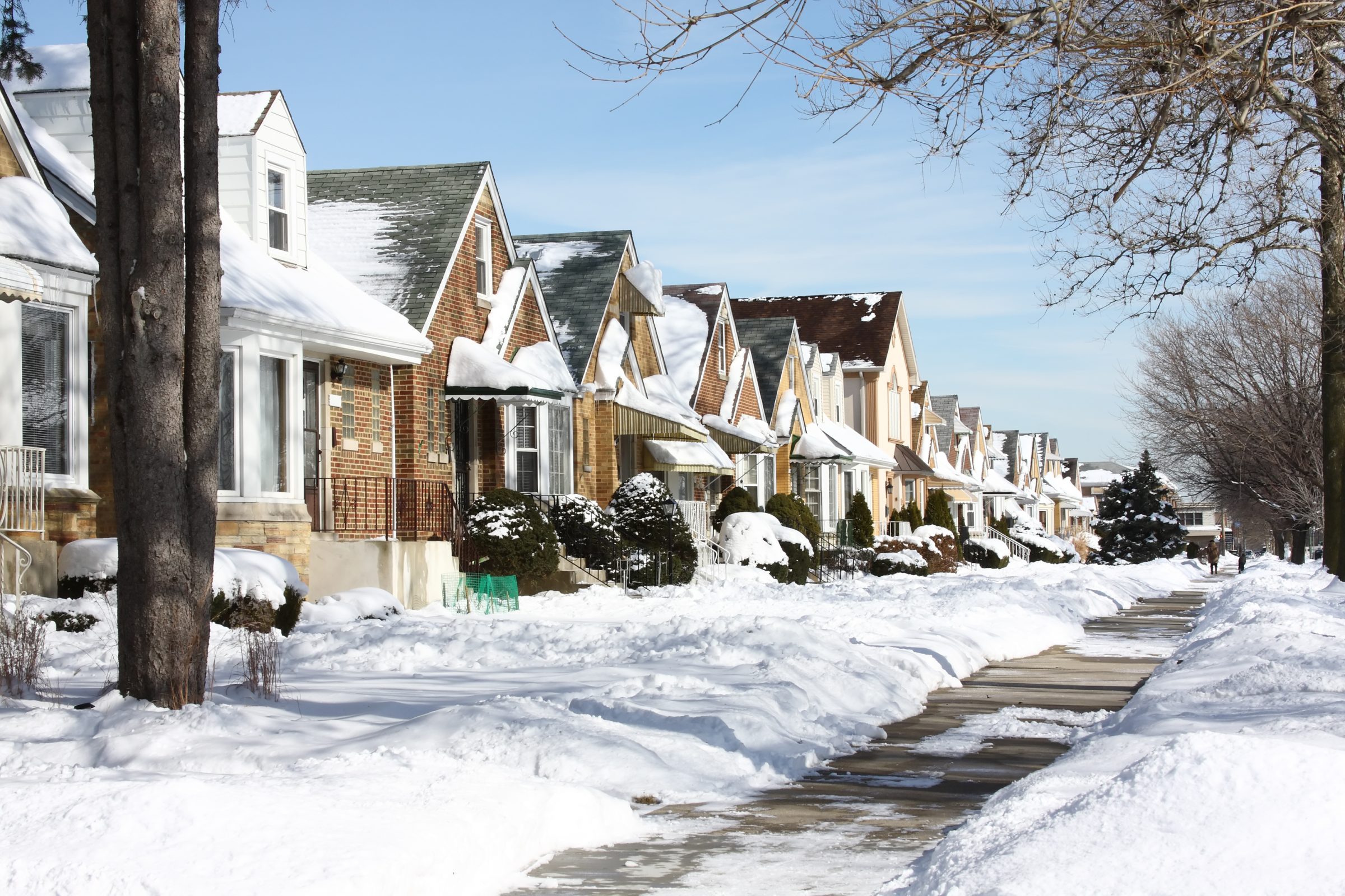 Snow-covered residential street with brick houses, snow-laden trees, and a clear blue sky. Sidewalks are partially shoveled, creating a peaceful winter scene.