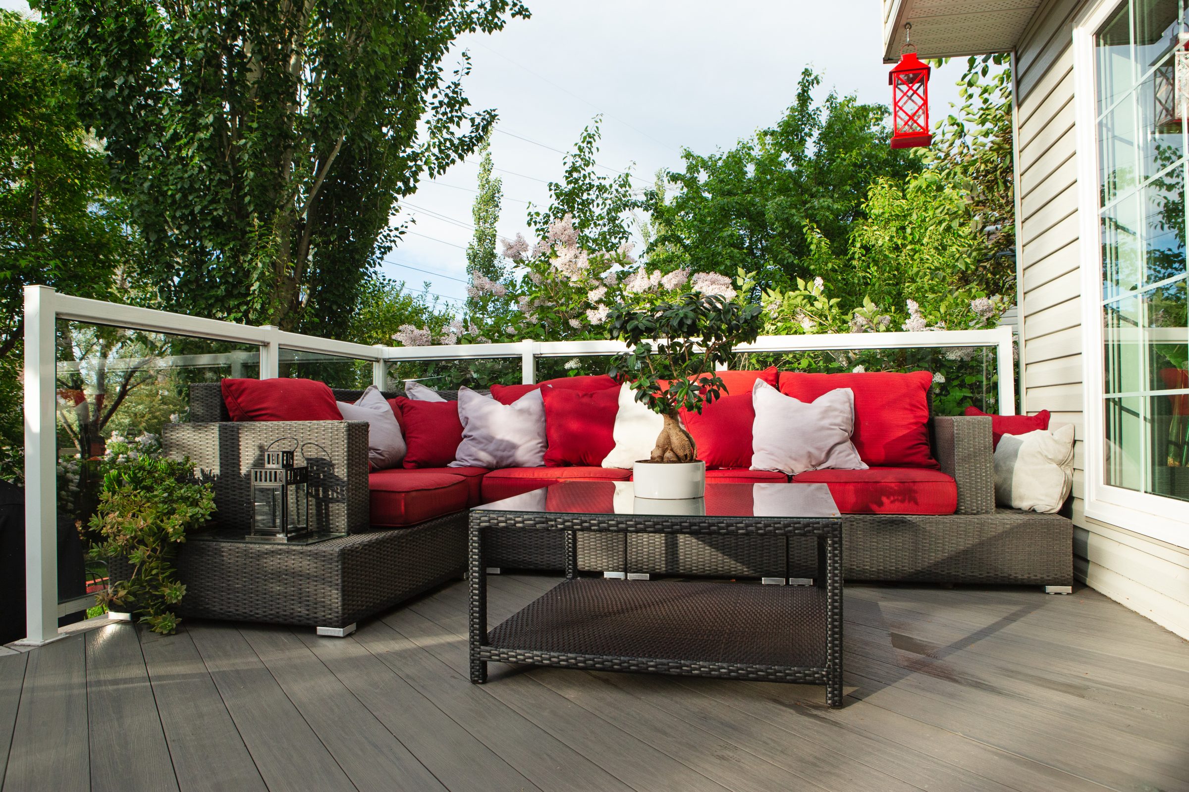 Outdoor patio with red cushions and pillows on wicker furniture. Lanterns and potted plant decorate the space, surrounded by greenery and trees.