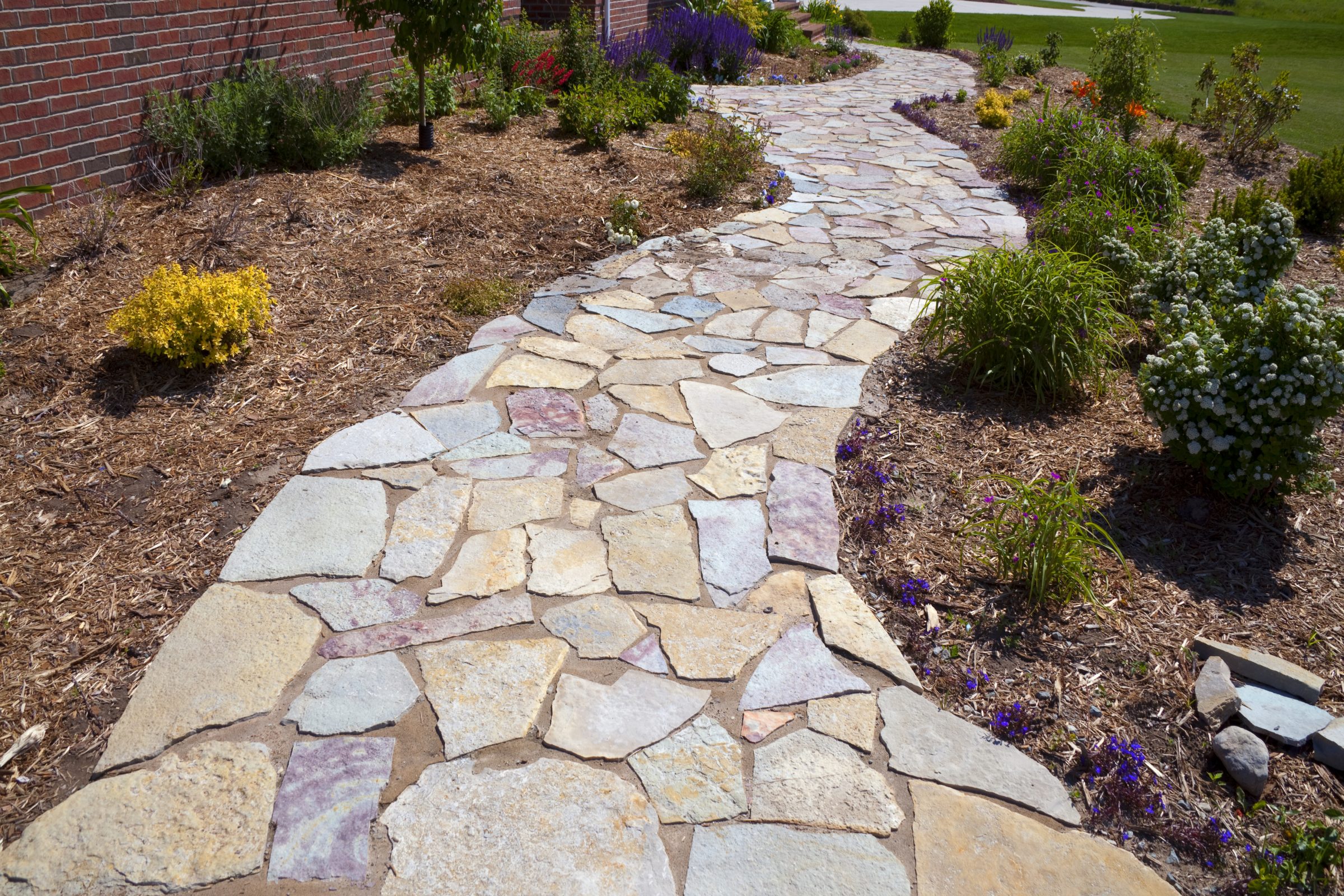 A curved stone path bordered by colorful flowers and greenery runs alongside a brick wall, under a clear sky.