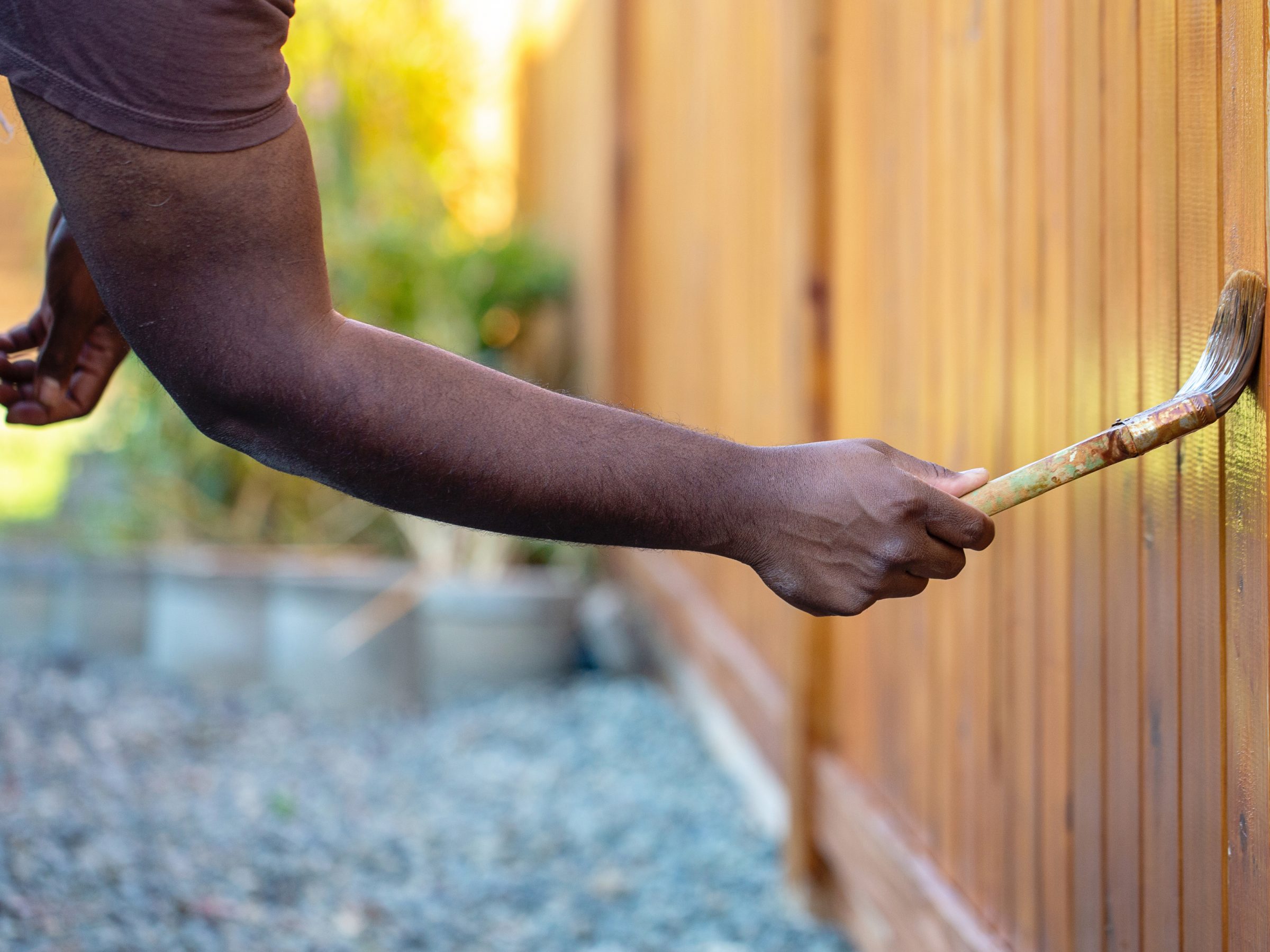 A person paints a wooden fence with a brush outdoors, surrounded by greenery and stones, focusing on their hand and the paintbrush.