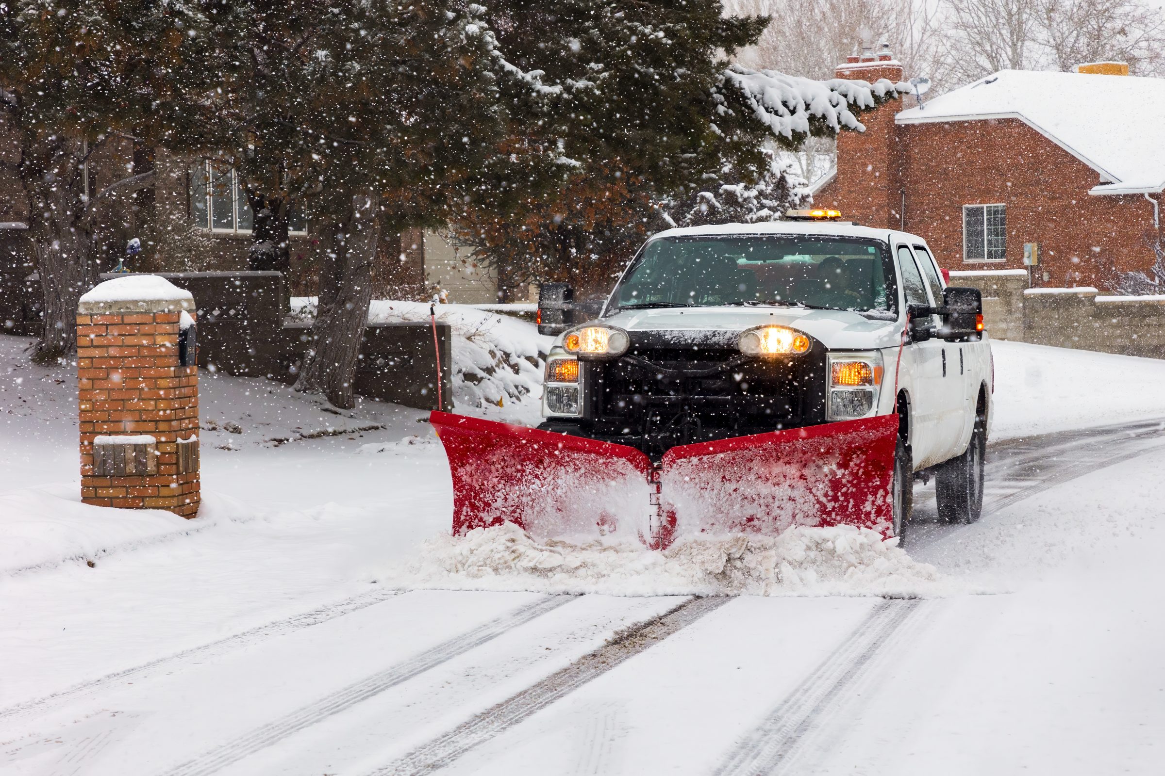 A snowplow clears a snowy residential street during snowfall. Brick houses and trees are covered in snow, creating a wintery neighborhood scene.