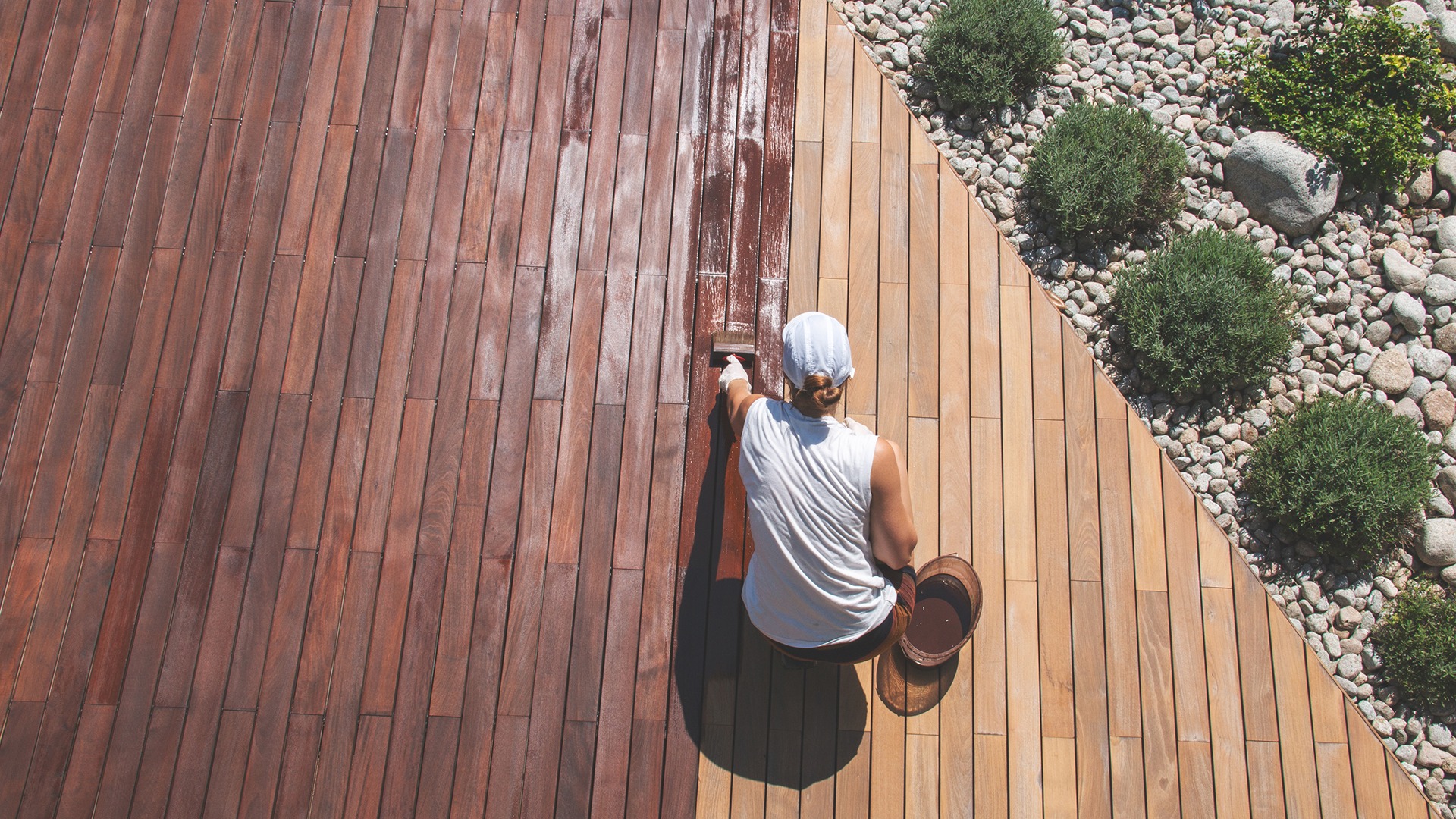 A person paints wooden decking, dividing stained and unstained areas, near a pebble and shrub garden.