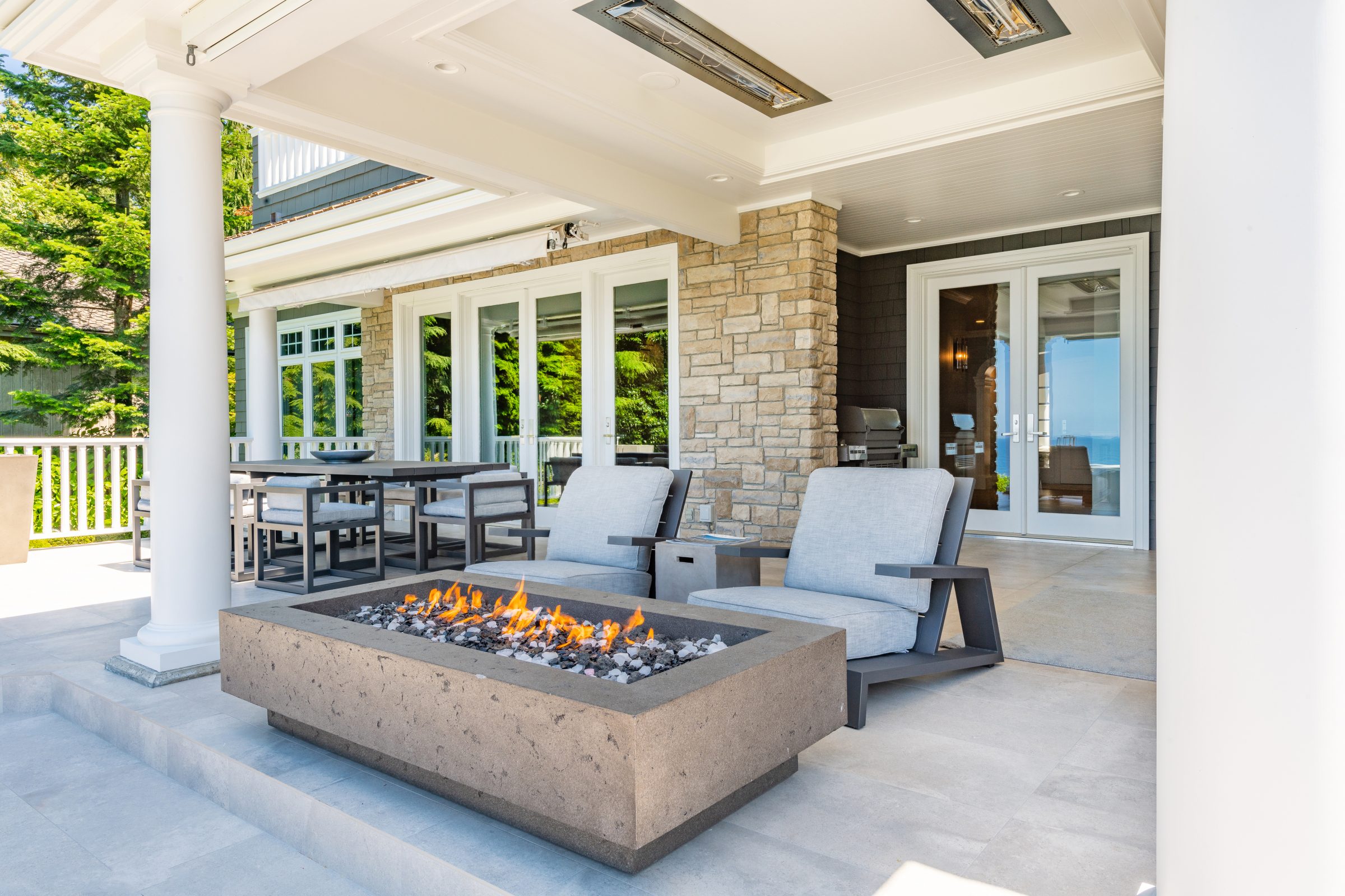 Outdoor patio with modern fire pit, comfy seating, dining table, and stone wall. Glass doors lead to interior. Scenic greenery surrounds.