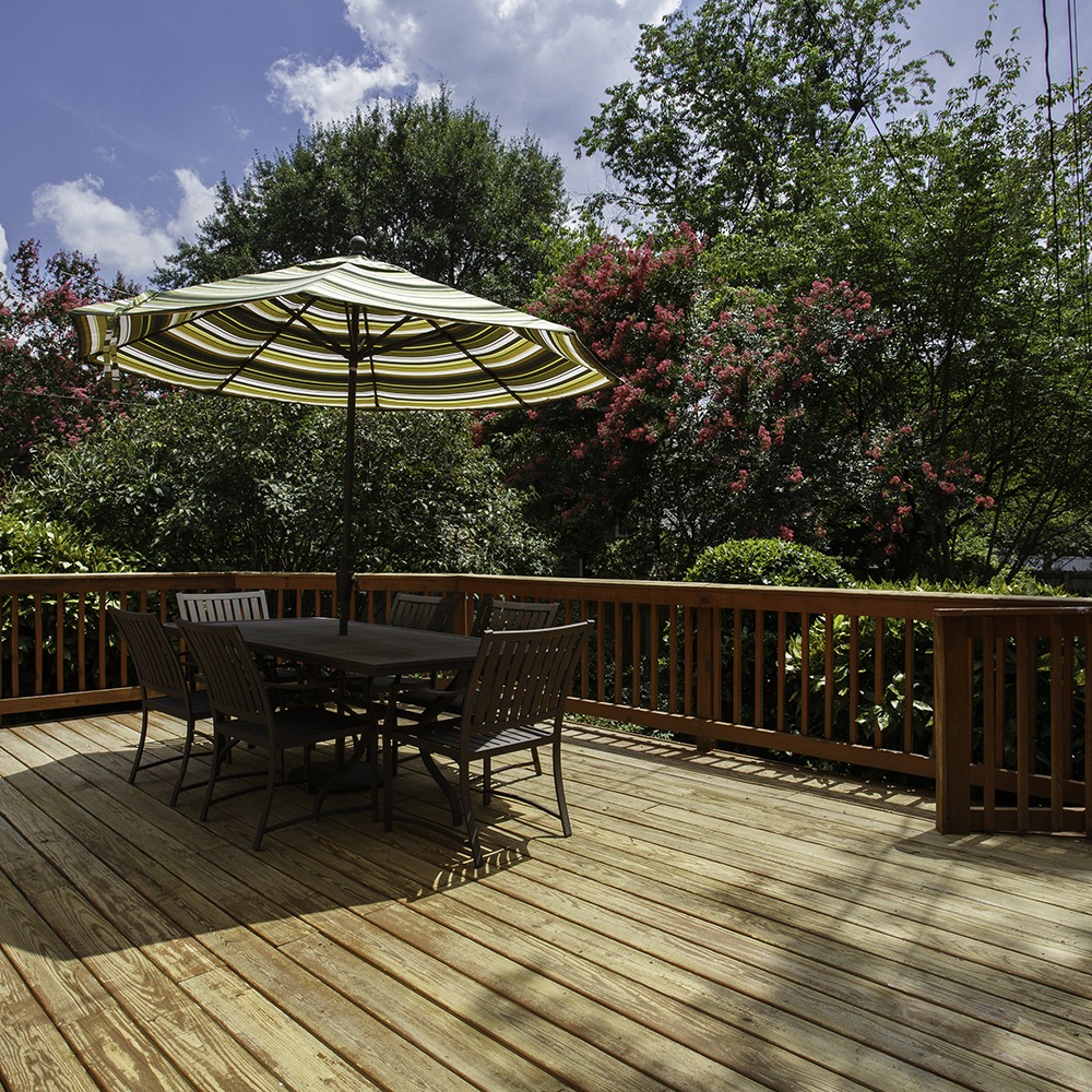 A wooden deck with a table, chairs, and a striped umbrella. Surrounded by lush greenery and trees under a partly cloudy sky.