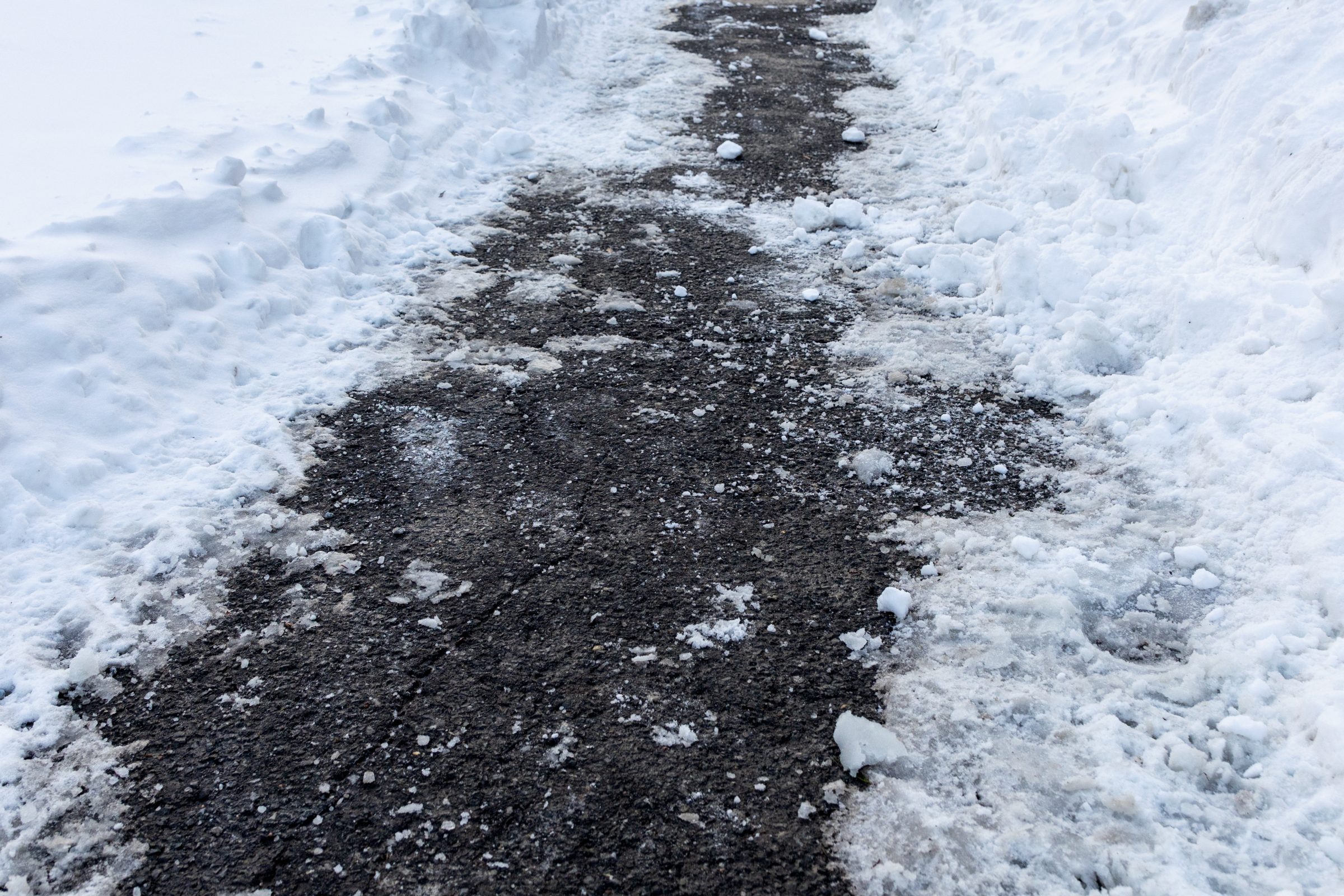 A snow-covered sidewalk with scattered ice, partially cleared, creating a narrow, icy path amid winter surroundings. No landmarks or people visible.