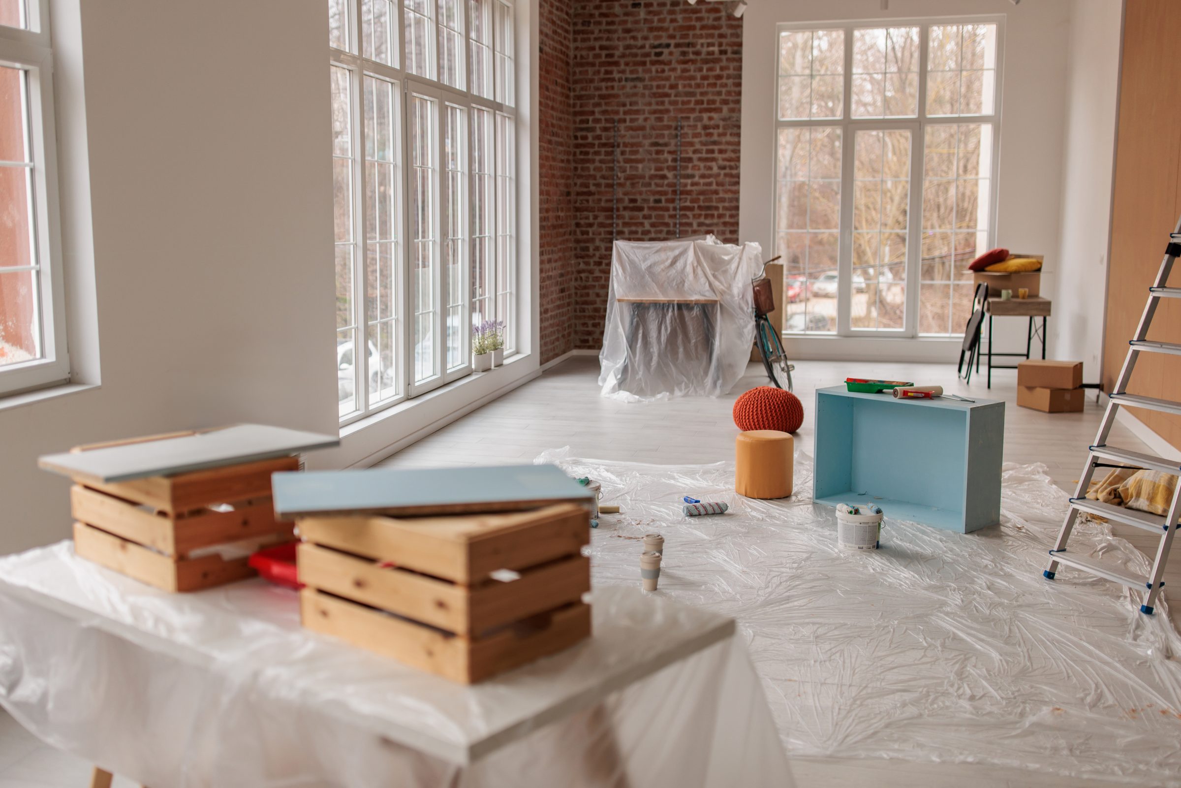Spacious room with large windows, brick walls, and scattered furniture for renovation. Painting supplies and ladder visible, creating a creative workspace atmosphere.