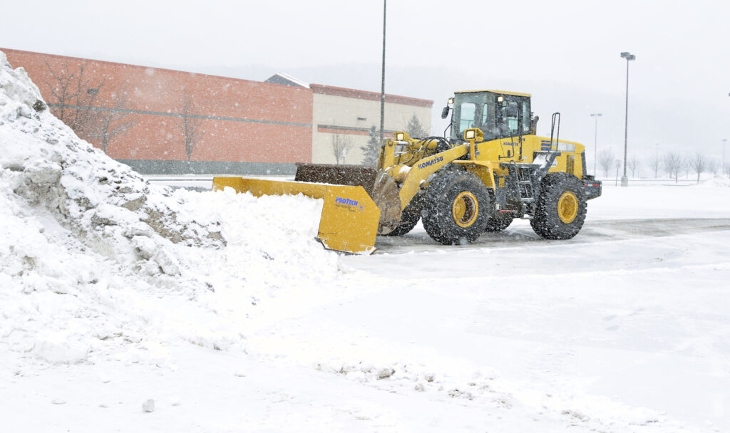 A yellow snowplow clears snow in a parking lot during snowfall, with a large brick building in the background.