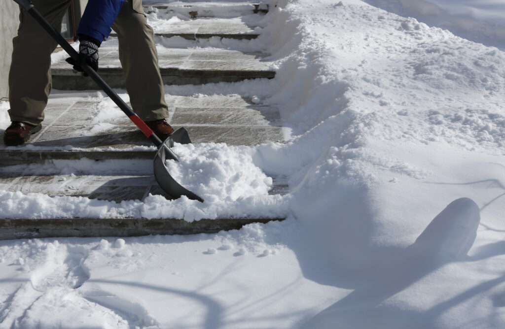 A person uses a shovel to clear snow from outdoor stairs. Snow is piled on both sides, indicating recent snowfall and a sunny day.