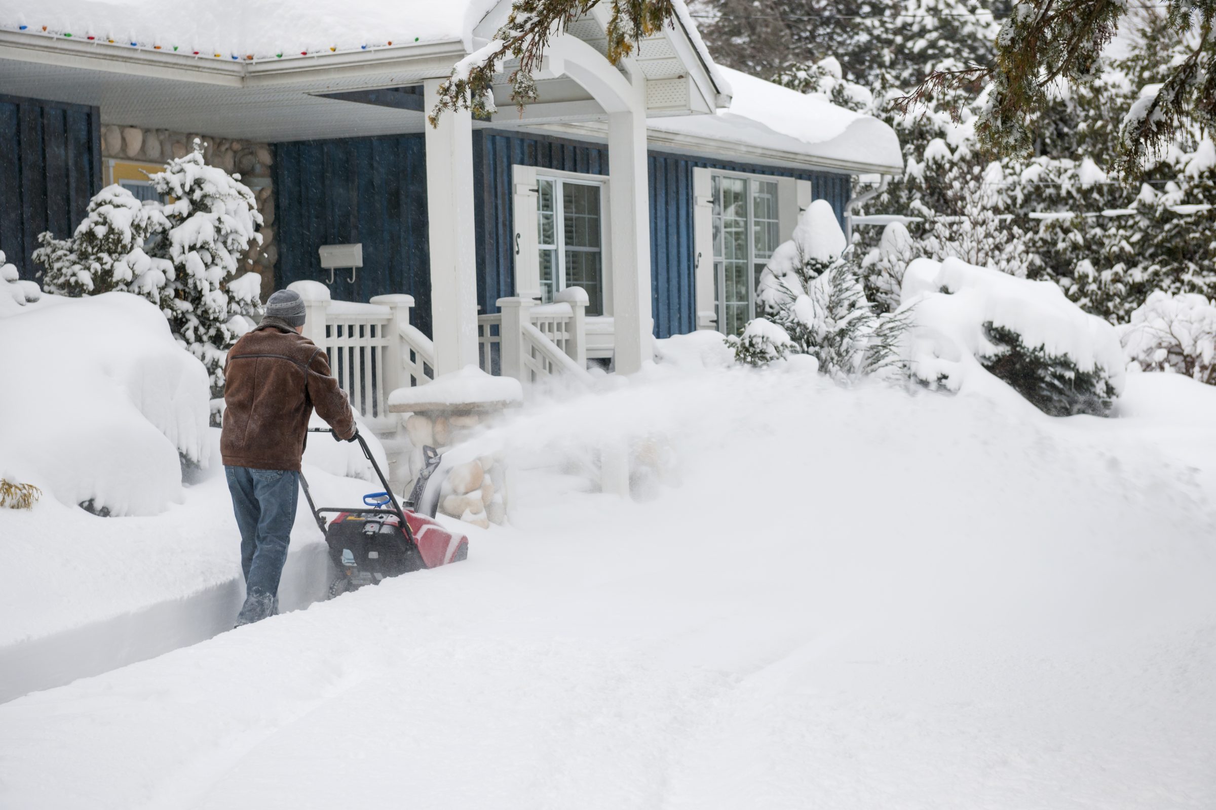 A person operates a snowblower on a snowy pathway outside a blue house with a porch and decorative lights.