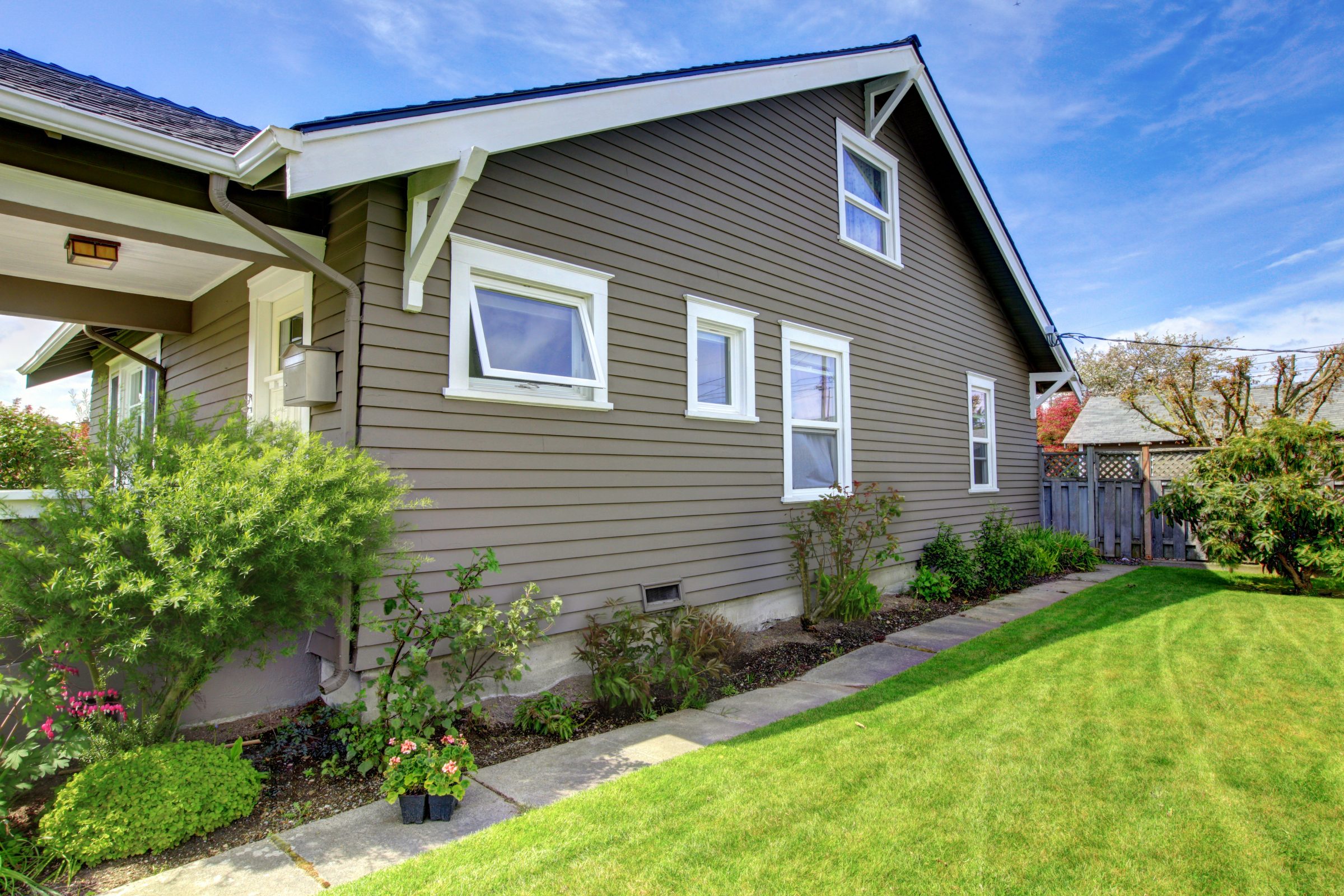 A well-maintained house with brown siding, white-trimmed windows, surrounded by a lush green lawn and garden, under a clear blue sky.