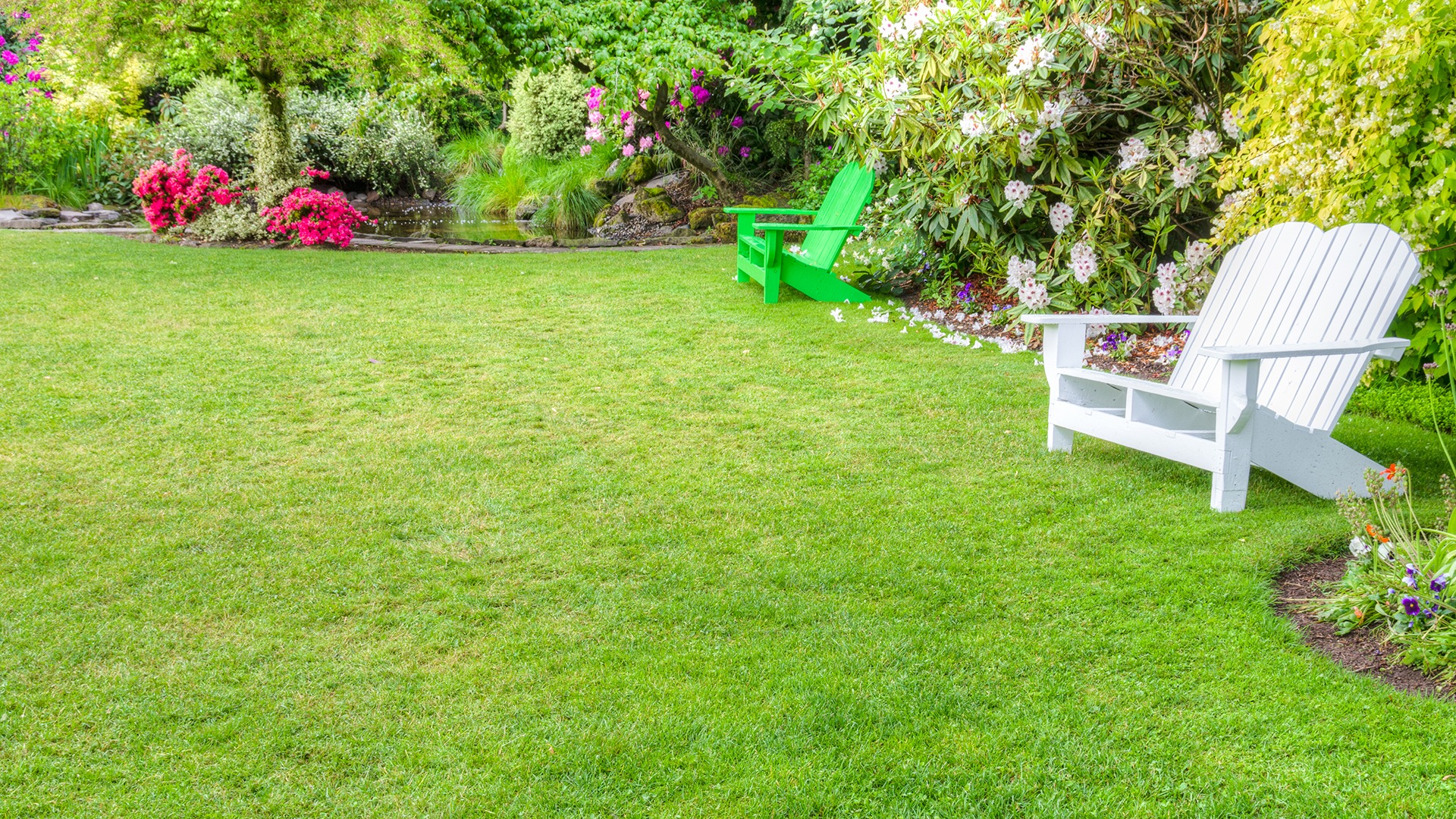 A lush garden features vibrant flowers, two Adirondack chairs, and a serene pond, creating a peaceful, inviting outdoor setting.