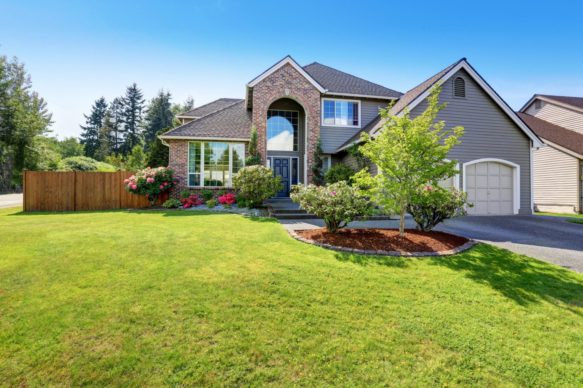 A two-story suburban house with a brick façade, manicured lawn, colorful flower beds, and a single garage, surrounded by lush trees under clear skies.