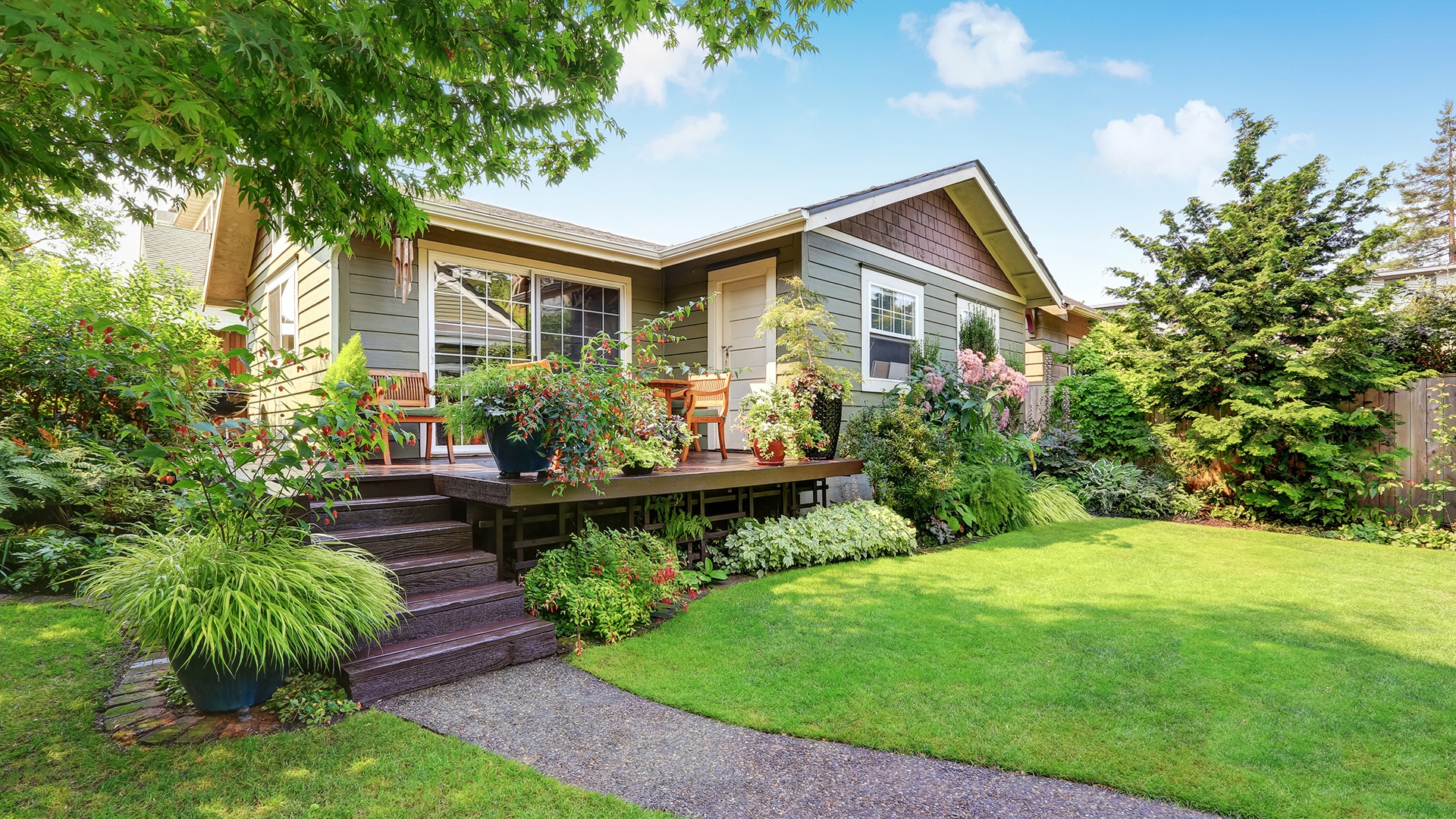 A charming house with a wooden porch, surrounded by lush green plants and trees, sits under a vibrant blue sky.