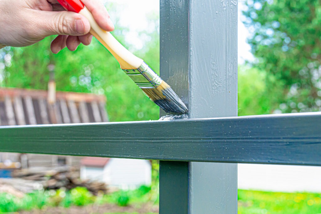A person paints an outdoor metal fence with a brush, surrounded by lush greenery and blurred background structures.