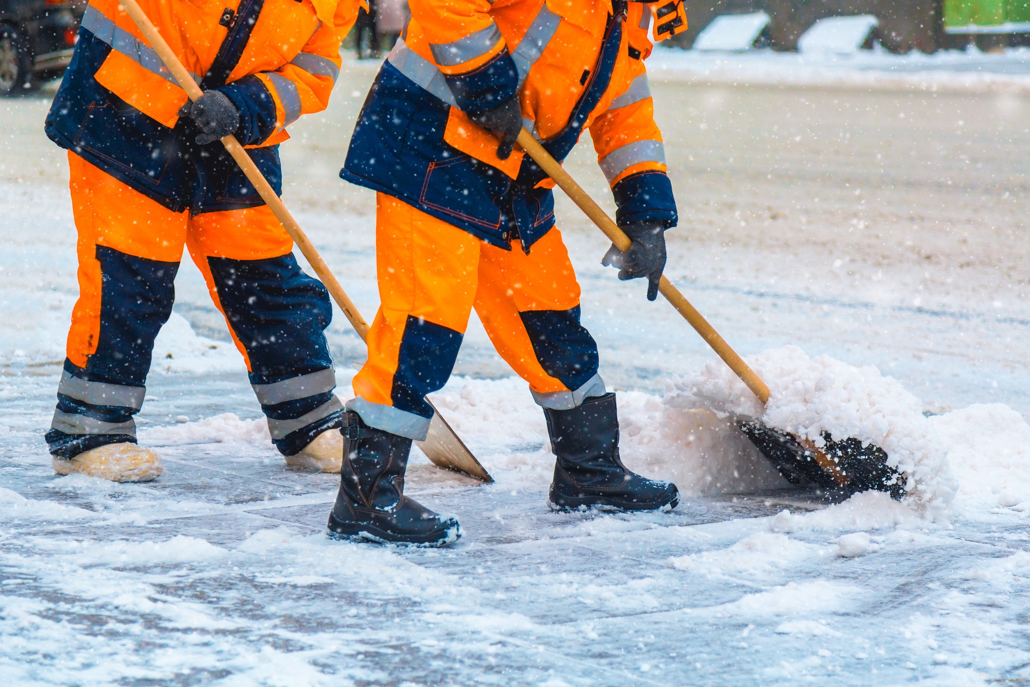 Two workers in orange safety gear shoveling snow from a paved surface during a snowfall
