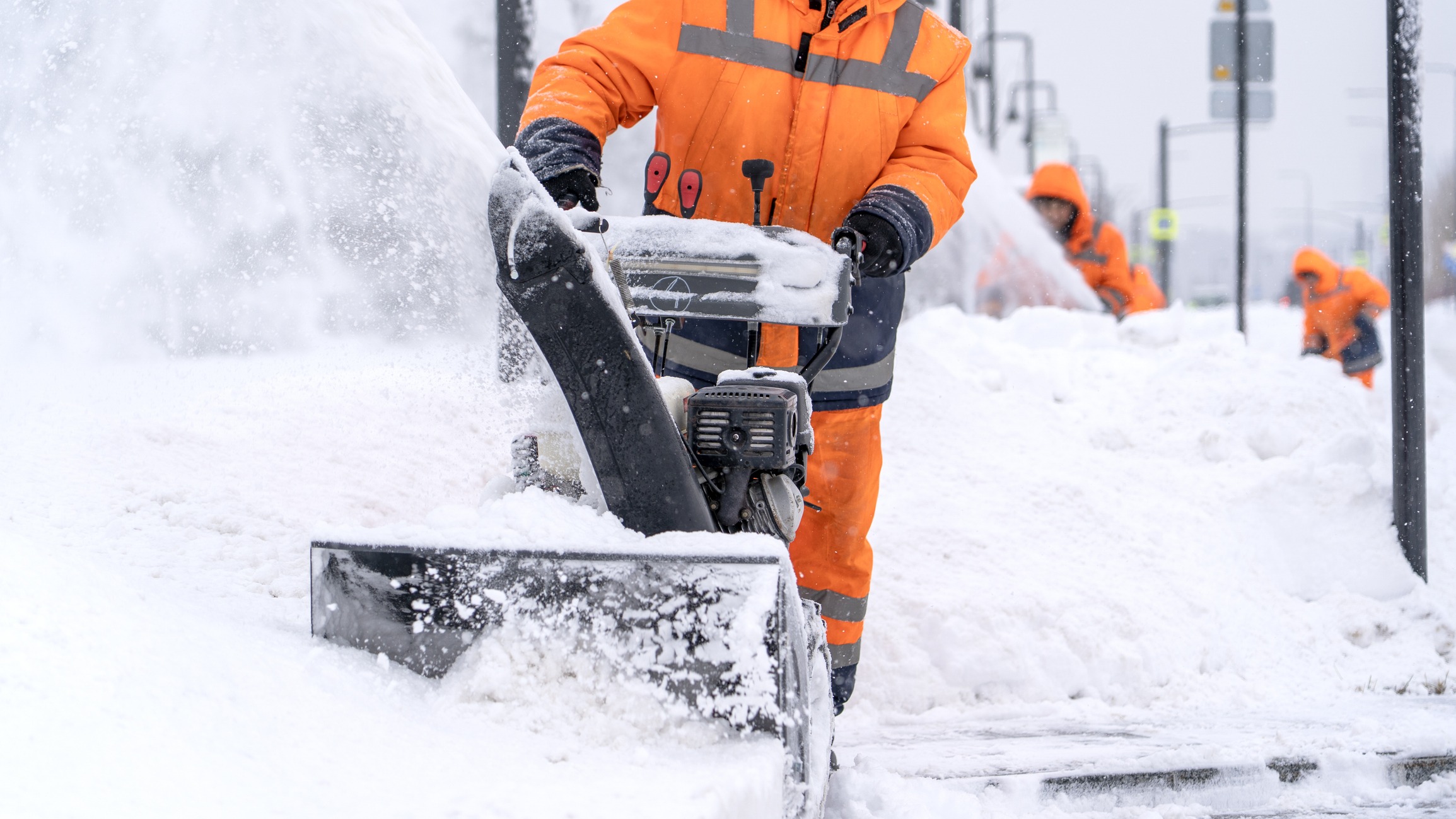 Worker in orange safety gear operating a snow blower on a snowy sidewalk